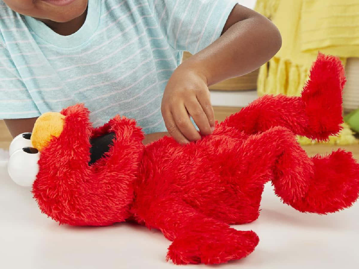 Elmo 桌布 壁紙 — 一個孩子輕輕觸摸放在白色表面上的亮紅色 Elmo 毛絨玩具。
