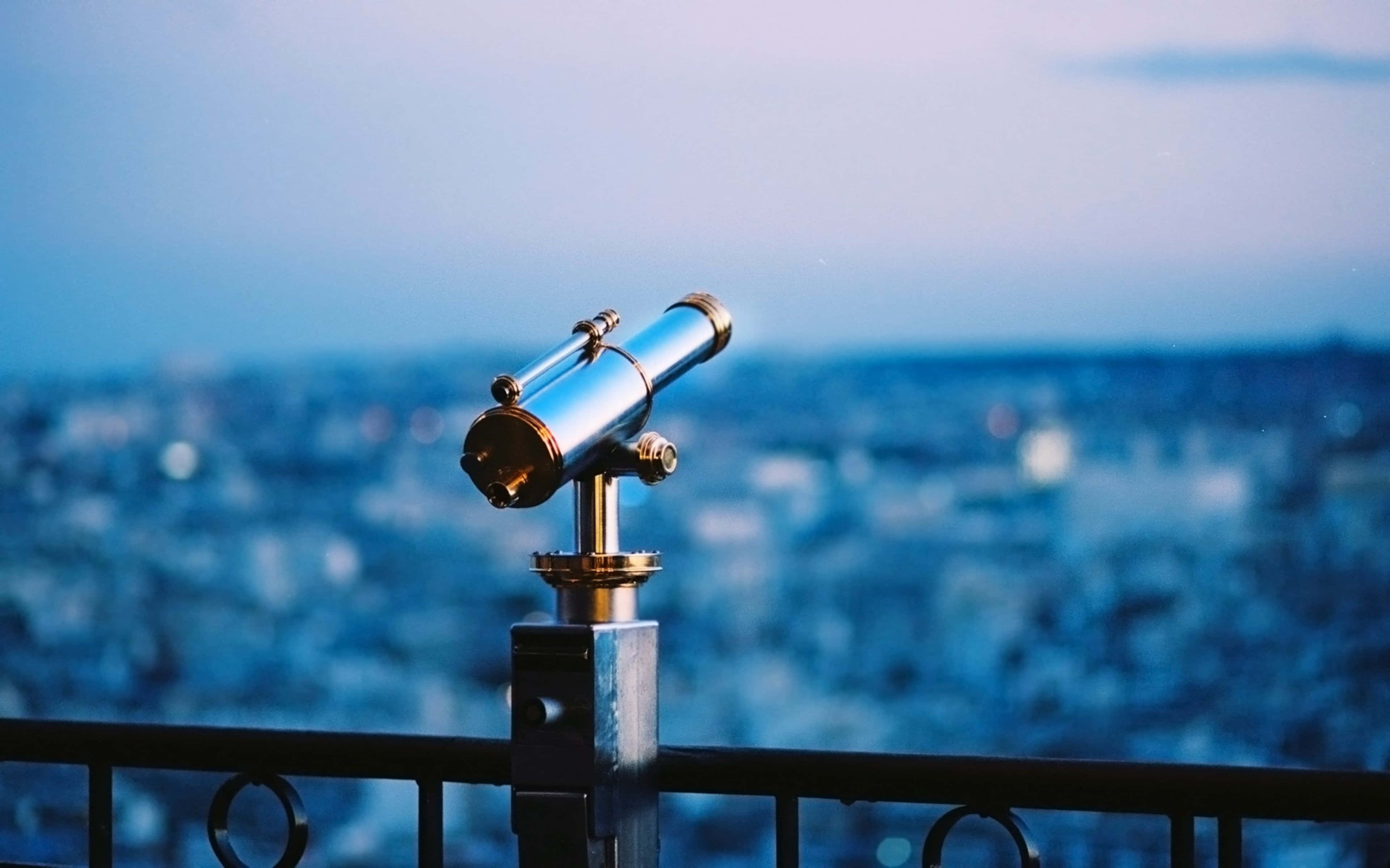 astronomy 桌布 壁紙 — 一個安裝在欄杆上的復古黃銅和鉻望遠鏡，俯瞰著柔和藍色暮色天空下模糊的城市景觀。