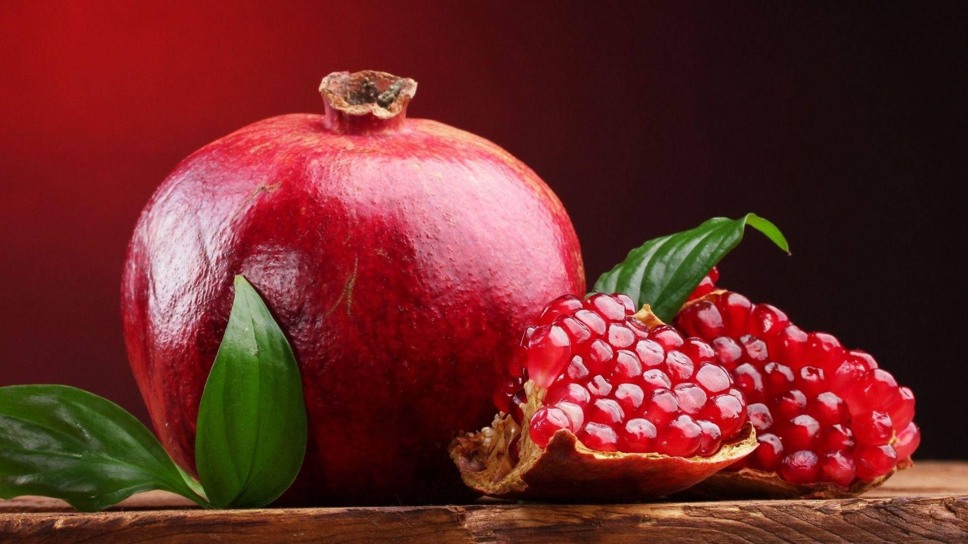 Fruit 桌布 壁紙 — 一個完整和切半的石榴，帶有多汁的籽和綠葉，放在質樸的木質表面上，背景是深紅色。