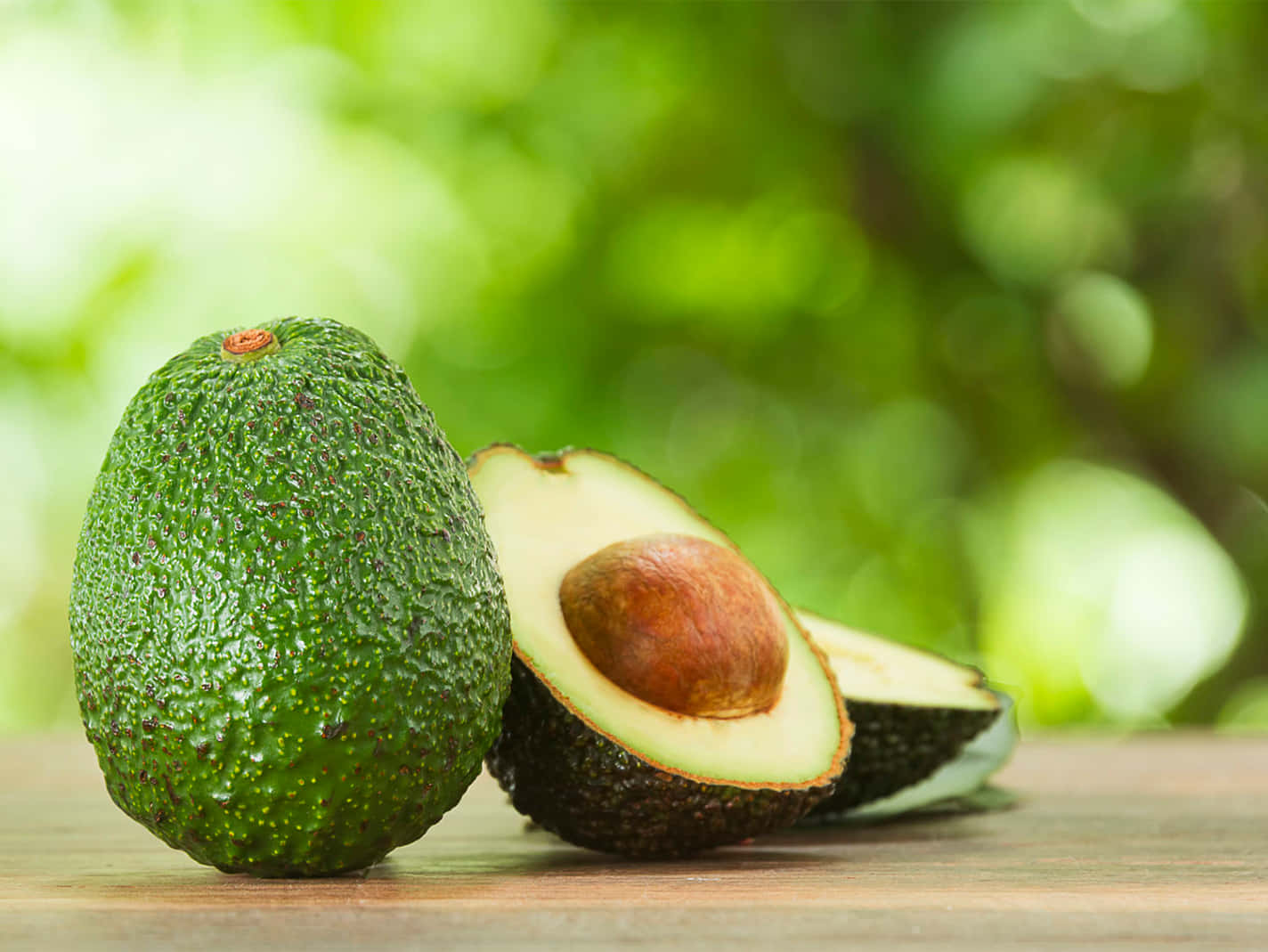 Avocado 桌布 壁紙 — 一個完整和切半的酪梨，帶有棕色果核，放在木質表面上，背景是柔和的綠色模糊效果。