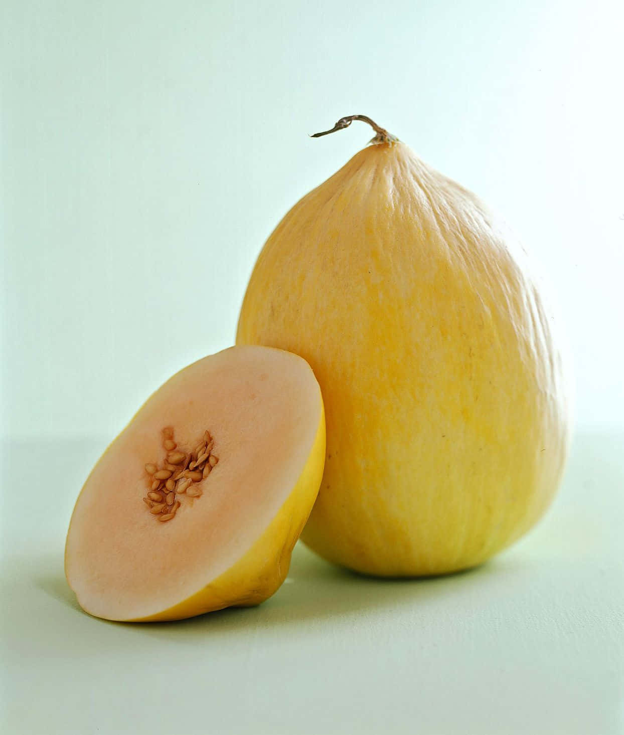 Fruit 桌布 壁紙 — 一個完整和切半的黃色哈密瓜，種子外露，放在淡綠色表面上，在柔和的工作室燈光下。