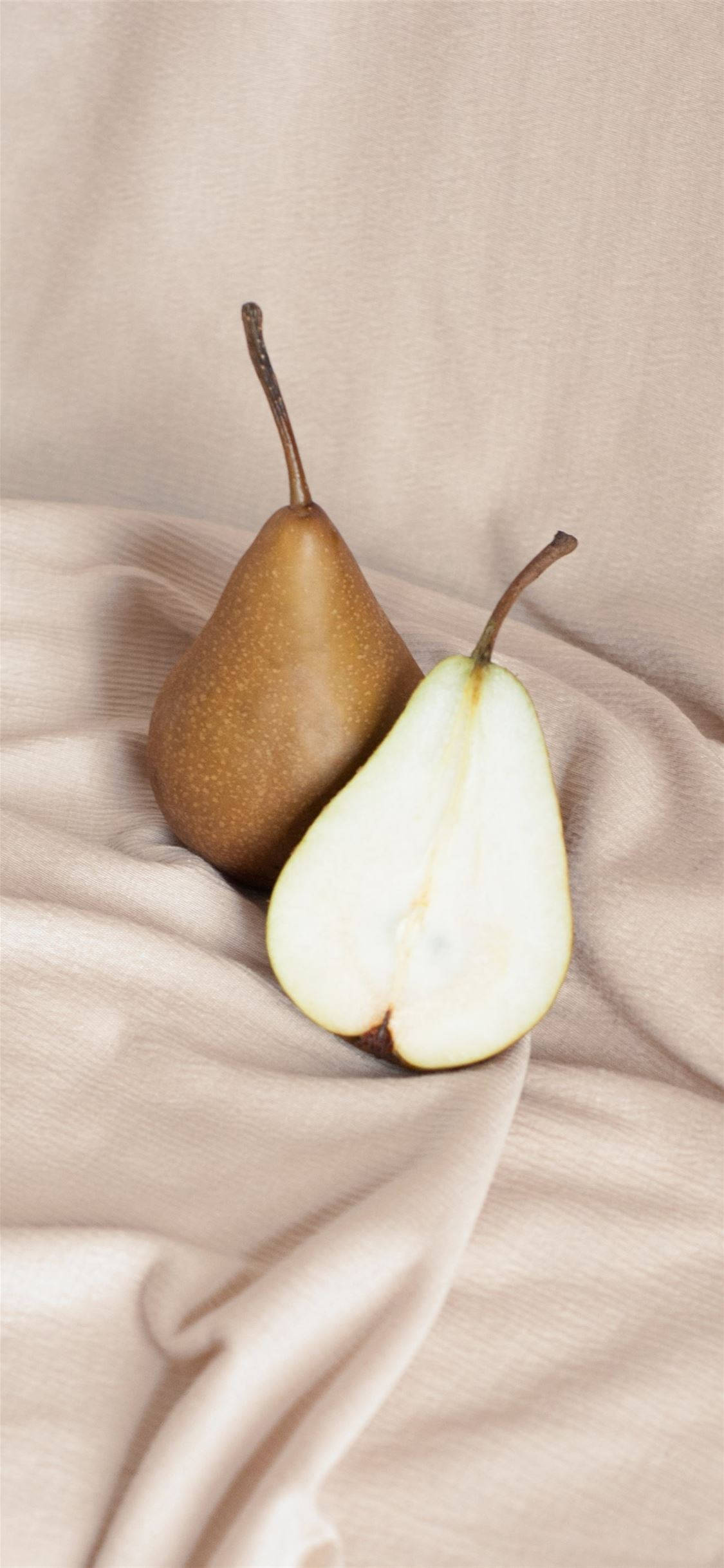 Fruits 桌布 壁紙 — 一個完整和半切的梨子放在柔軟的米色布料上，帶有輕柔的褶皺和自然光。