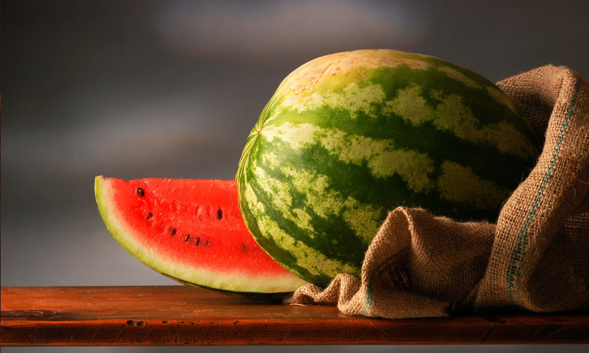 Watermelon 桌布 壁紙 — 一個完整的西瓜和多汁的一片放在木桌上，旁邊有一個麻布袋，背景是深色漸變。