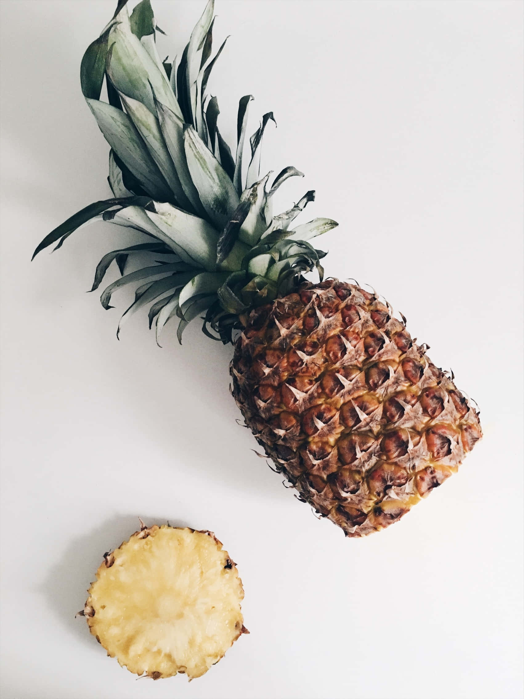 Pineapple 桌布 壁紙 — 一個完整的鳳梨，帶有綠色尖刺葉子，以及一個切片橫截面，在純白色背景上露出多汁的黃色果肉。
