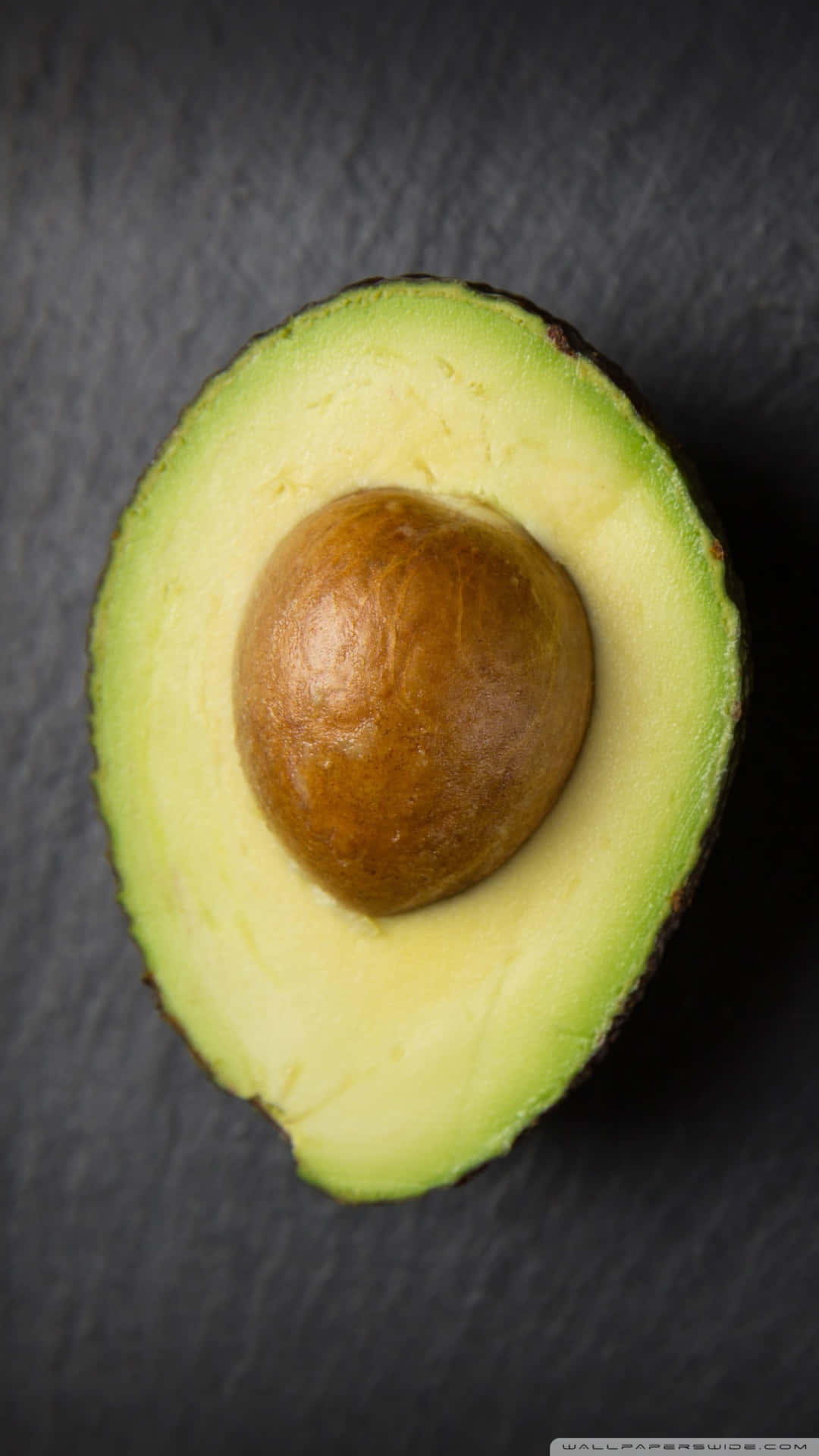Avocado iPhone 桌布 壁紙 — 一個完美切半的酪梨，帶有奶油狀的綠色果肉和棕色果核，放在深色板岩表面上。