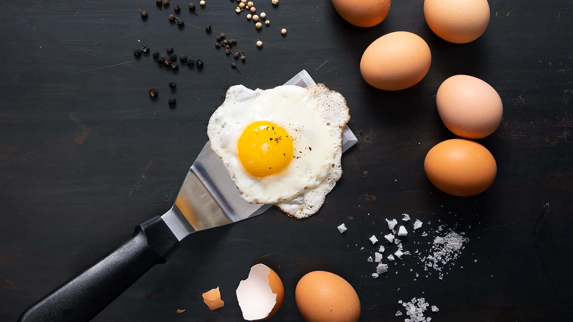 Egg White 桌布 壁紙 — 一個完美煎好的太陽蛋放在鍋鏟上，周圍有全蛋、鹽和胡椒，置於深色鄉村風格的表面上。