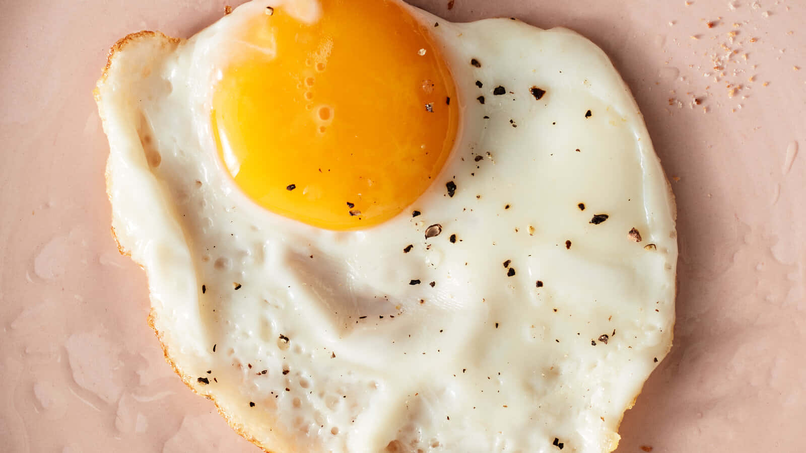 Egg 桌布 壁紙 — 一個完美的煎蛋，帶有鮮黃色的蛋黃和點綴的黑胡椒，放在柔軟的粉紅色表面上。