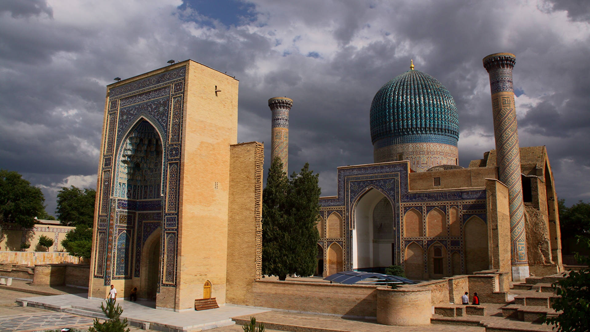 Uzbekistan 桌布 壁紙 — 一個宏偉的伊斯蘭建築群，擁有綠松石瓷磚圓頂和宣禮塔，背景是戲劇性的多雲天空。