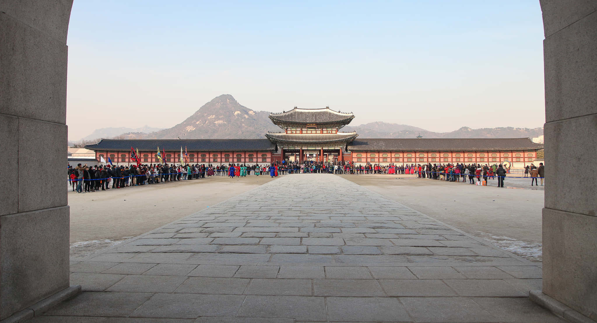 韓國 桌布 壁紙 — 一個宏偉的傳統韓國宮殿庭院，以石拱門為框架，遊客聚集在宏偉的大門前，背景是遠山。
