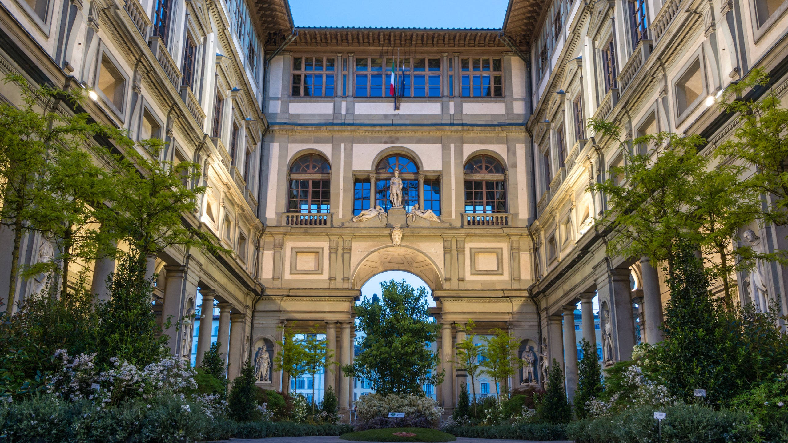 建築 桌布 壁紙 — 一個宏偉的古典庭院，擁有對稱的建築、拱形走道和晴朗藍天下的茂密綠植。