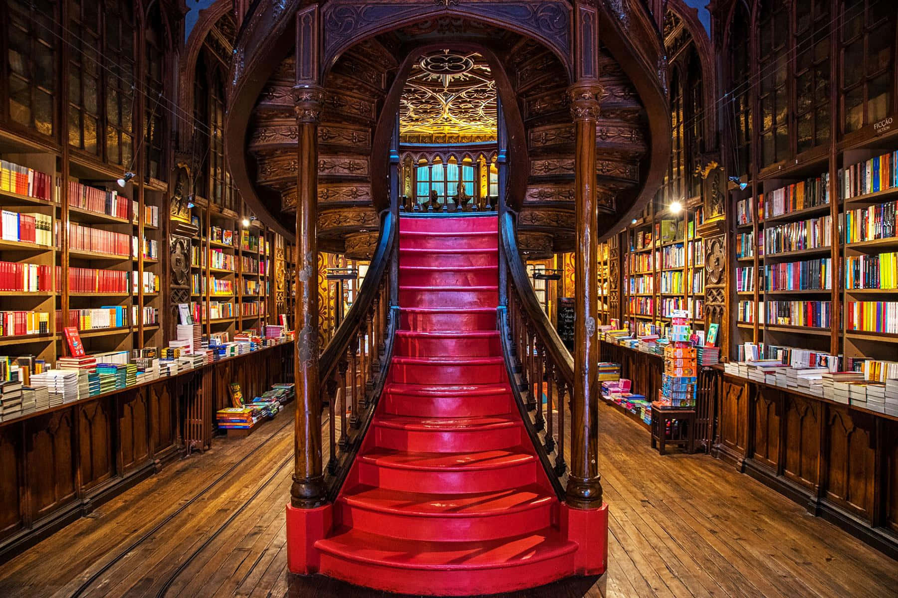 The Hogwarts Library 桌布 壁紙 — 一個宏偉的圖書館，擁有高聳的木製書架和一條引人注目的紅色地毯樓梯向上延伸。