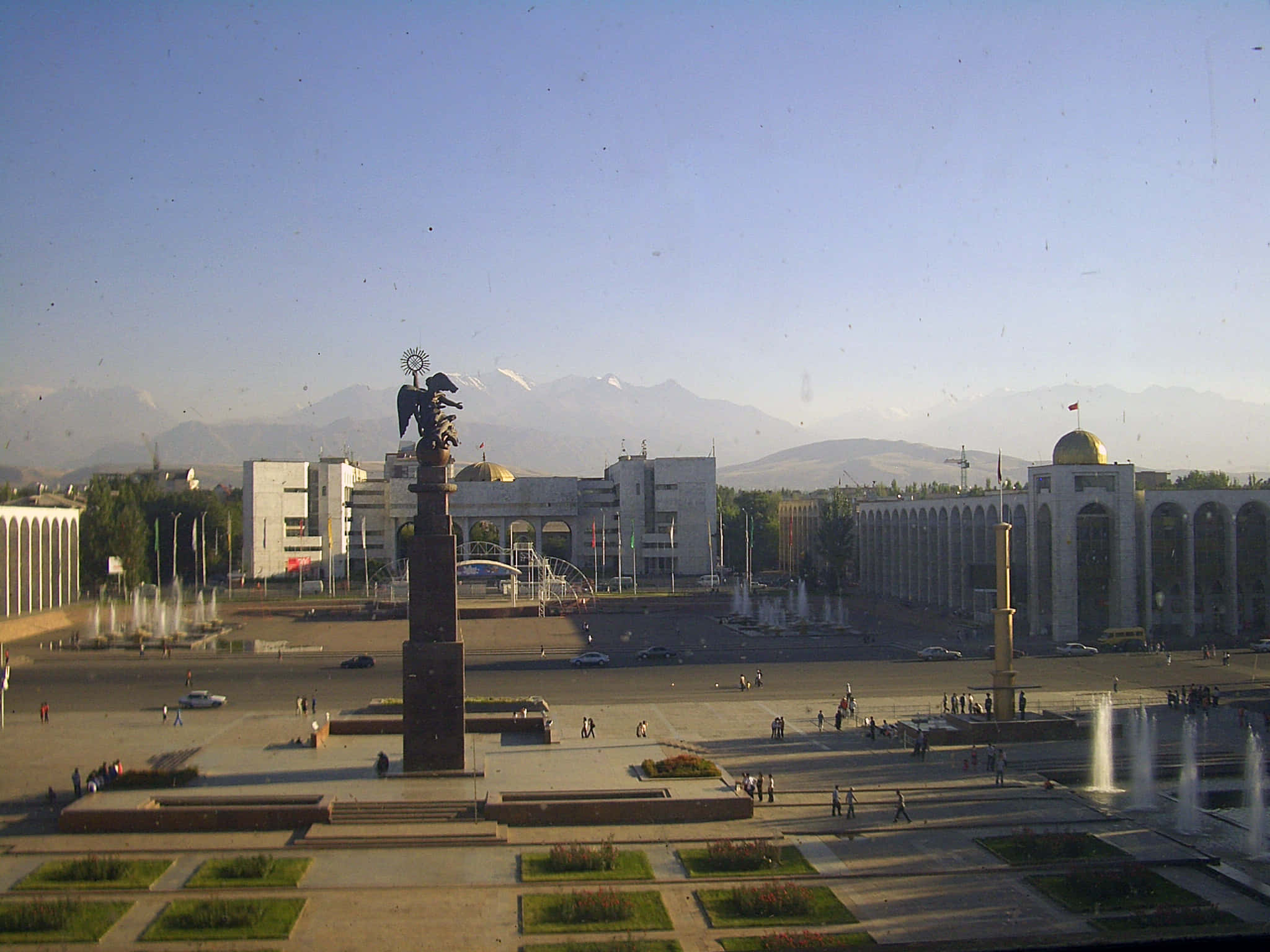 Kyrgyzstan 桌布 壁紙 — 一個宏偉的城市廣場中央有一座帶有翅膀雕塑的紀念碑，周圍是噴泉和帶有金色圓頂的現代建築，背景是白雪覆蓋的山脈，在清澈的藍天下。