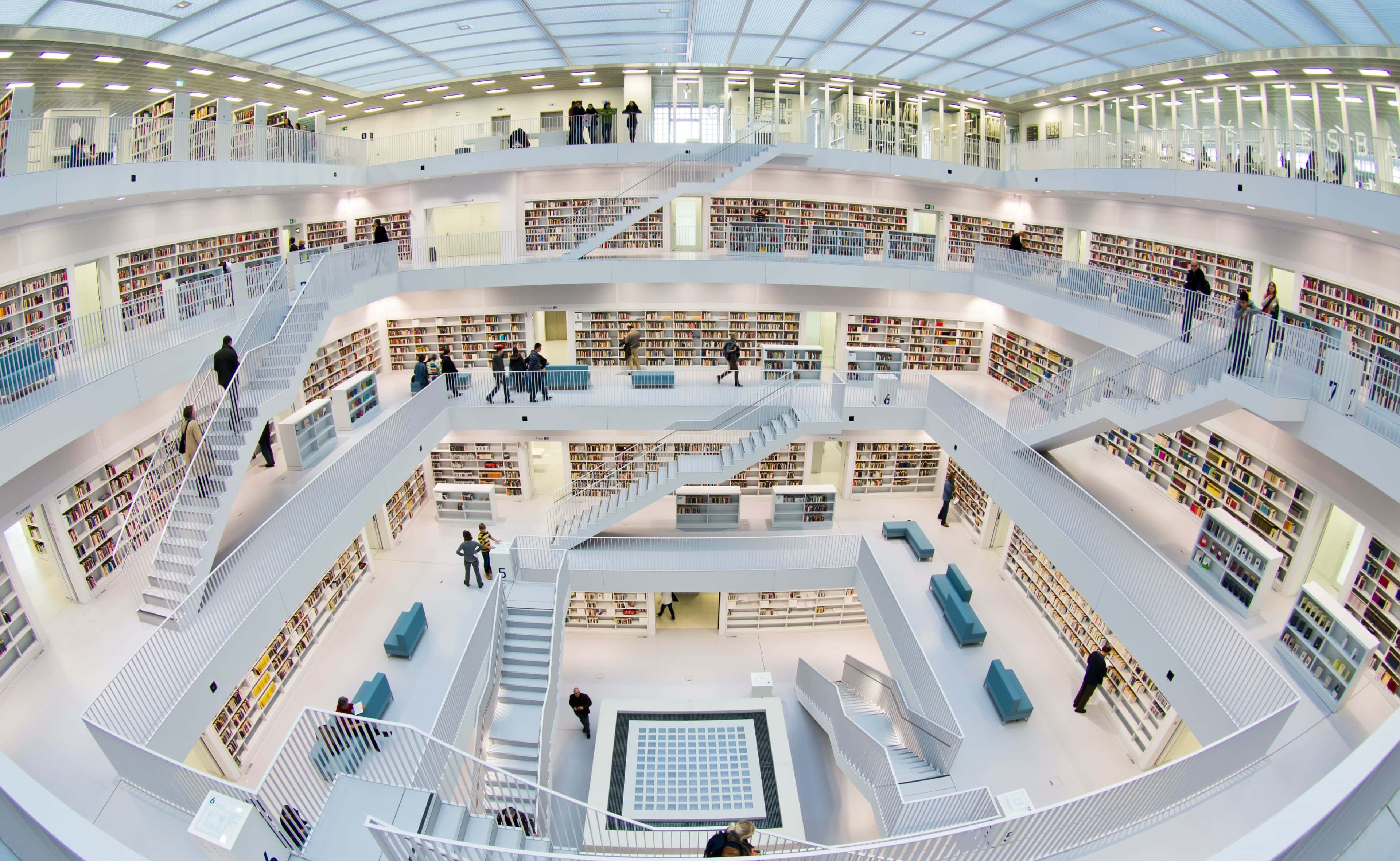 Europe 桌布 壁紙 — 一個宏偉的多層圖書館，設有白色螺旋樓梯和書架，書架上擺滿了色彩繽紛的書籍，上方有明亮的吊燈。