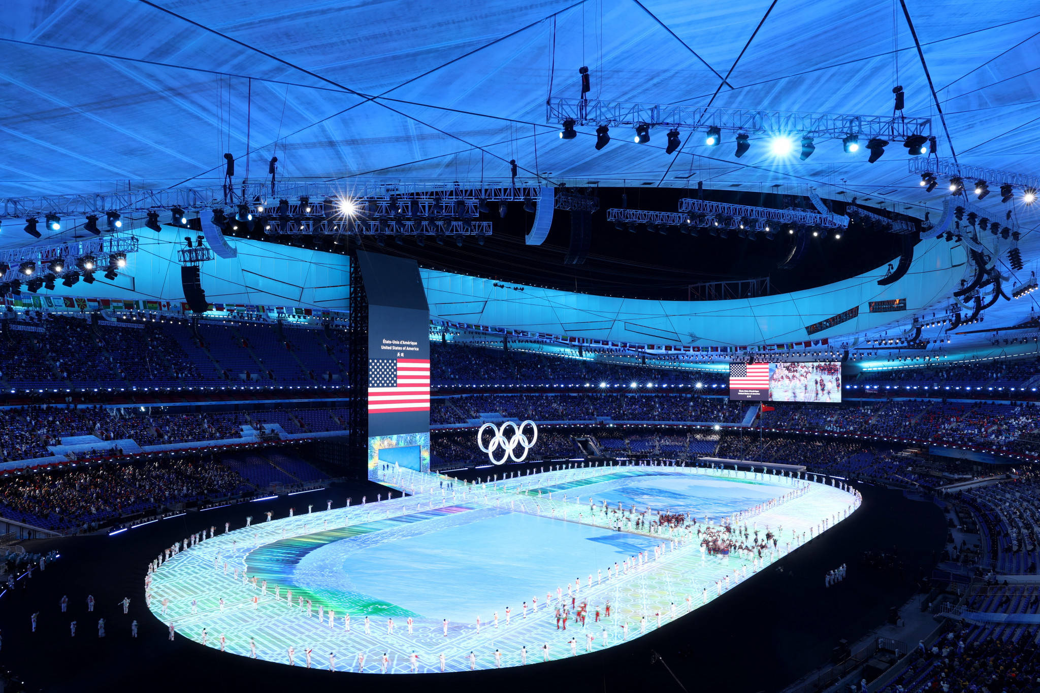 Winter Olympics 桌布 壁紙 — 一個宏偉的奧林匹克體育場在夜晚，發光的溜冰場被歡呼的人群包圍，上方是藍色照明的穹頂。