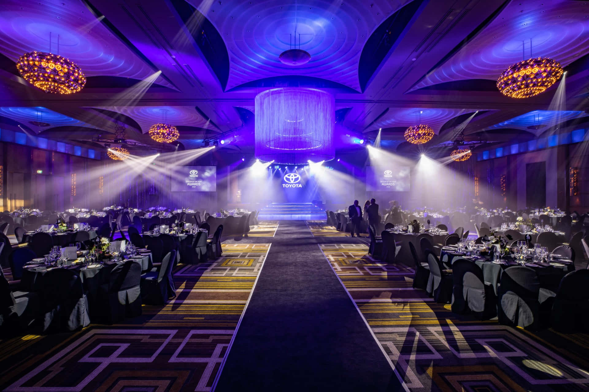 Corporate Event 桌布 壁紙 — 一個宏偉的宴會廳，沐浴在紫色和金色的燈光中，配有水晶吊燈和一個帶有豐田標誌的中央舞台。