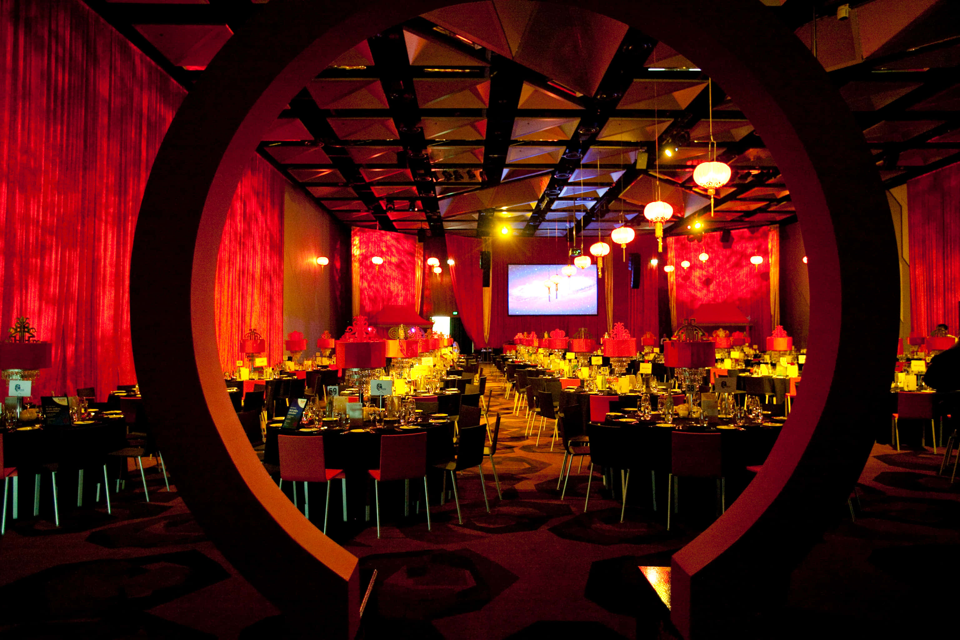 Black Tie Event 桌布 壁紙 — 一個宏偉的宴會廳沐浴在深紅色的燈光中，透過一個大圓形框架觀看，餐桌已為優雅的活動佈置好。