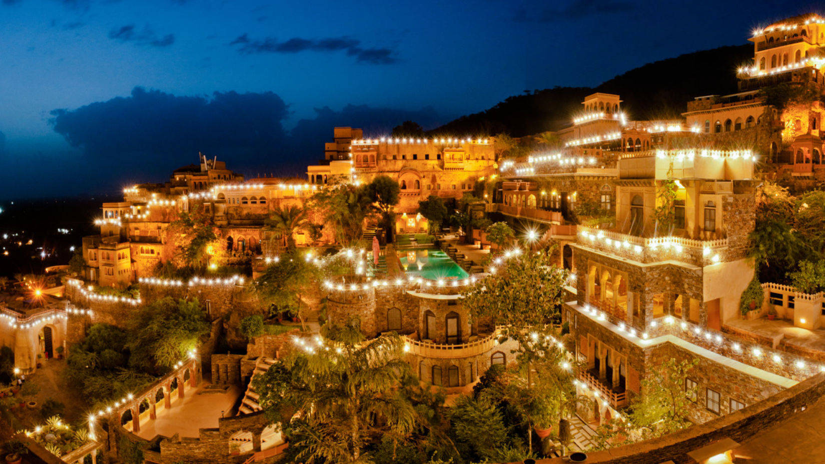 India 桌布 壁紙 — 一個宏偉的山坡度假村在夜晚溫暖地發光，串燈照亮了石頭建築和茂密的花園，在深藍色的天空下。