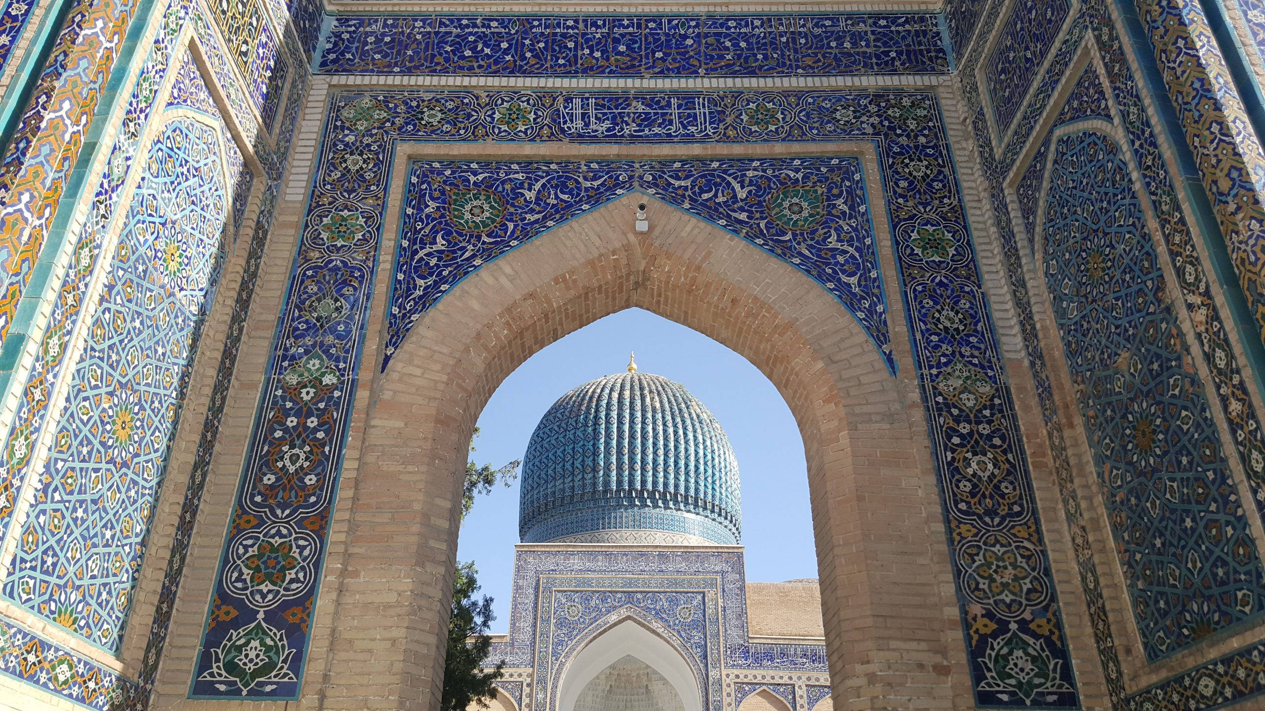 Uzbekistan 桌布 壁紙 — 一個宏偉的拱門，飾有精緻的藍色和金色瓷磚，在清澈的藍天下框住了一座雄偉的圓頂清真寺。