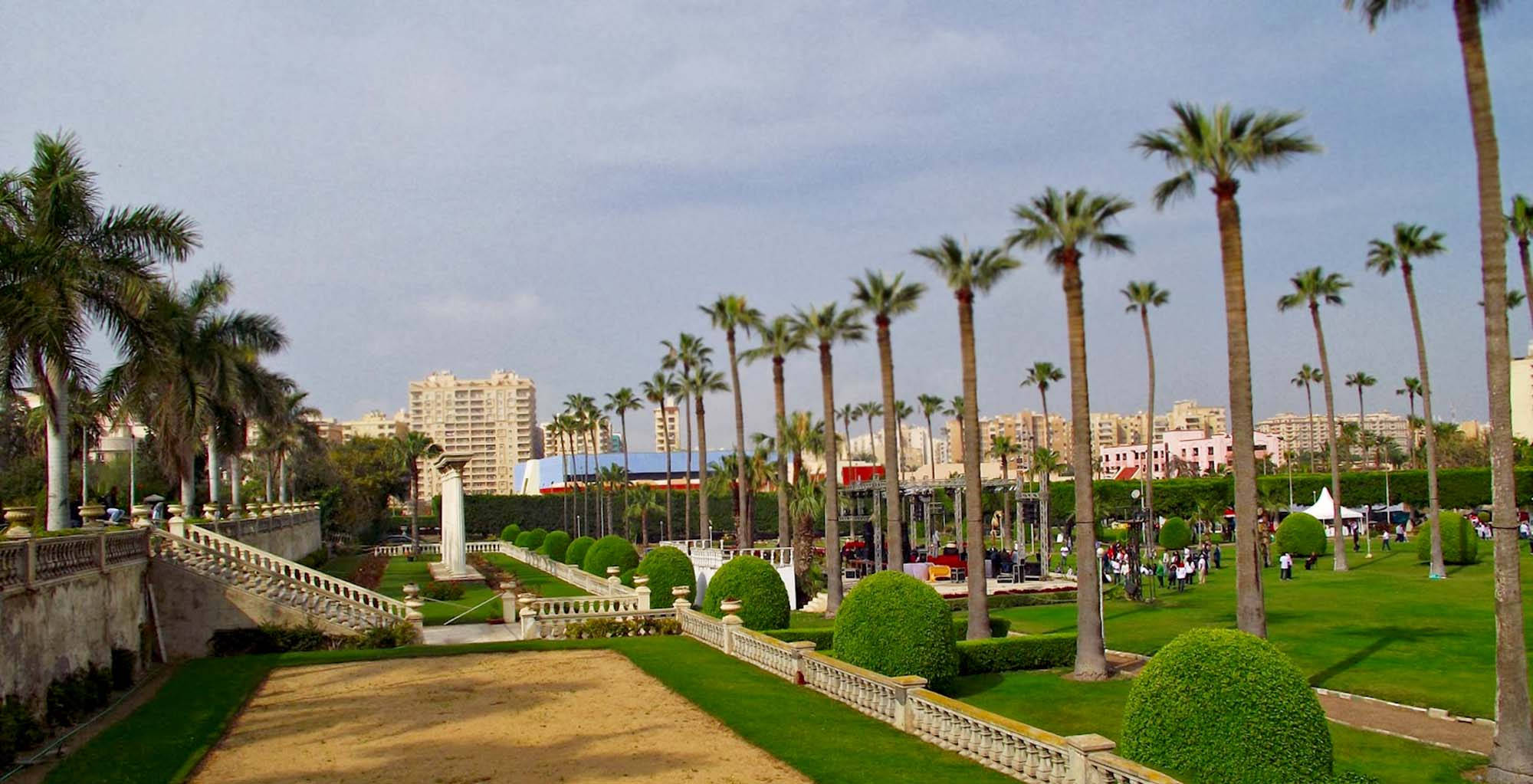 Alexandria 桌布 壁紙 — 一個宏偉的景觀花園，種有棕櫚樹、修剪整齊的樹籬和石欄杆，在晴朗的天空下延伸至城市天際線。
