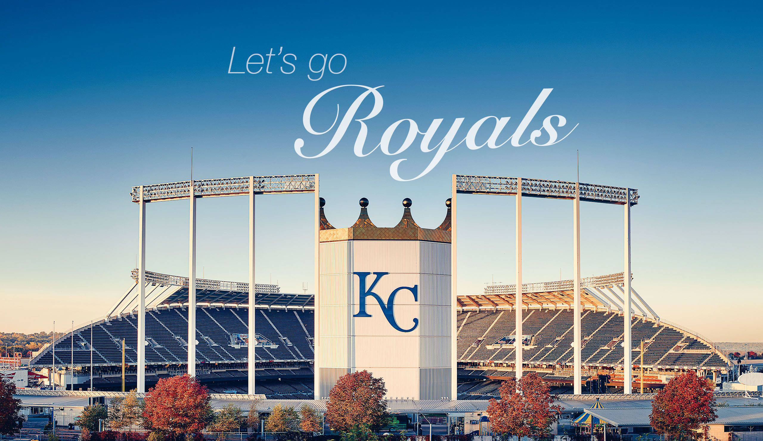 Kansas 桌布 壁紙 — 一個宏偉的棒球場，帶有 'KC Royals' 品牌標誌，背景是晴朗的藍天，前景有秋樹。