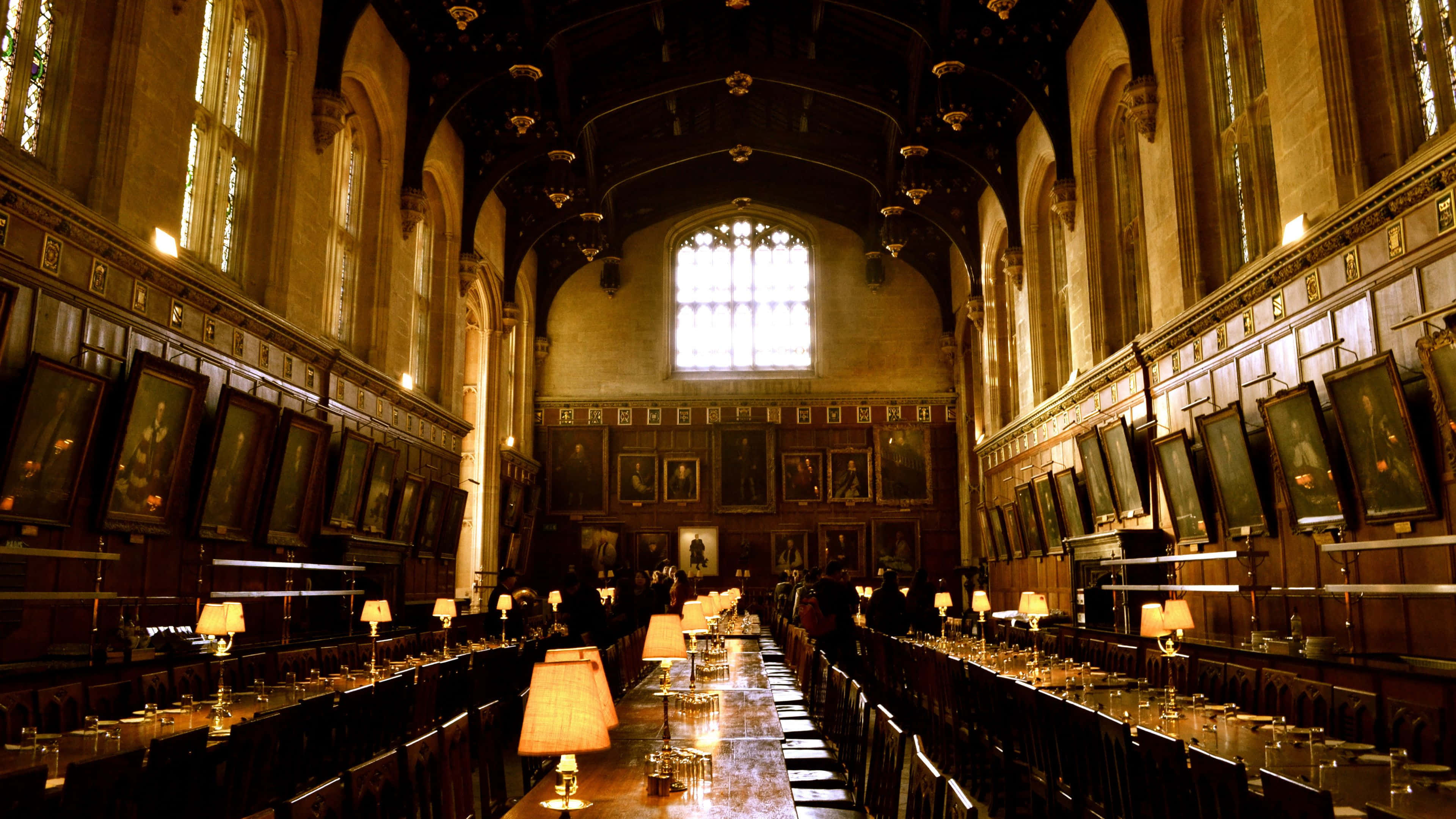 Great Hall Harry Potter Desktop 桌布 壁紙 — 一個宏偉的歷史宴會廳，配有長木桌、牆上掛滿肖像畫，以及來自燈具和彩繪玻璃窗的溫暖光線。