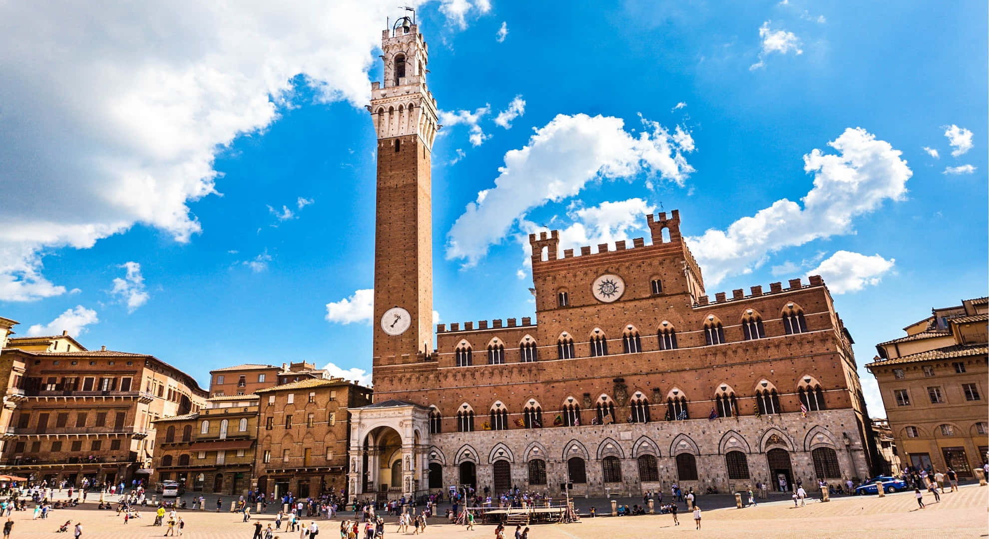 Piazza Del Campo 桌布 壁紙 — 一個宏偉的歷史廣場，擁有高聳的鐘樓和華麗的磚造建築，在明亮的藍天下有蓬鬆的白雲。