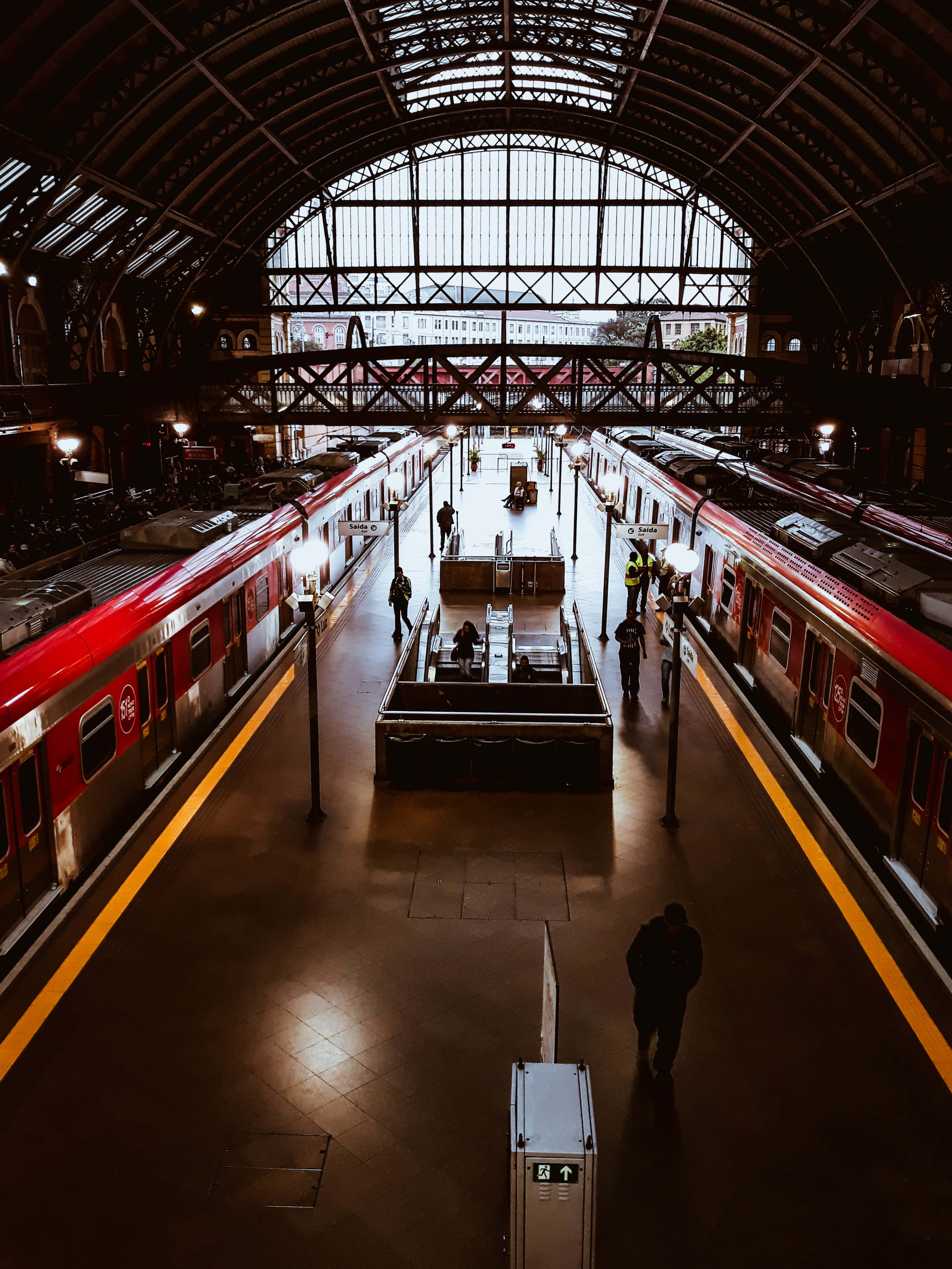 Train Station 桌布 壁紙 — 一個宏偉的火車站，擁有拱形玻璃天花板和紅色火車，月台兩側有旅客在溫暖的人造燈光下行走。