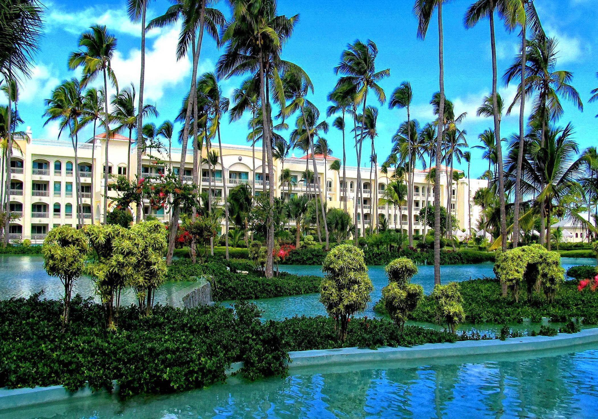 Hotel 桌布 壁紙 — 一個宏偉的熱帶度假酒店，擁有棕櫚樹和碧綠的泳池，在明亮的藍天下。