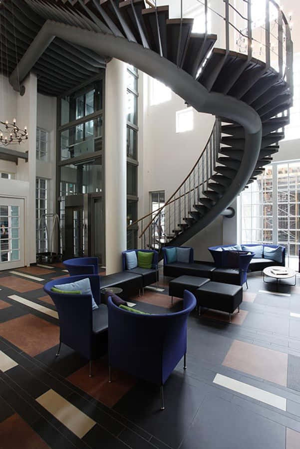 Interior Design 桌布 壁紙 — 一個宏偉的現代大廳設有華麗的螺旋樓梯，配有深色金屬踏板和玻璃欄杆，周圍環繞著深藍色休閒椅和圖案瓷磚地板。