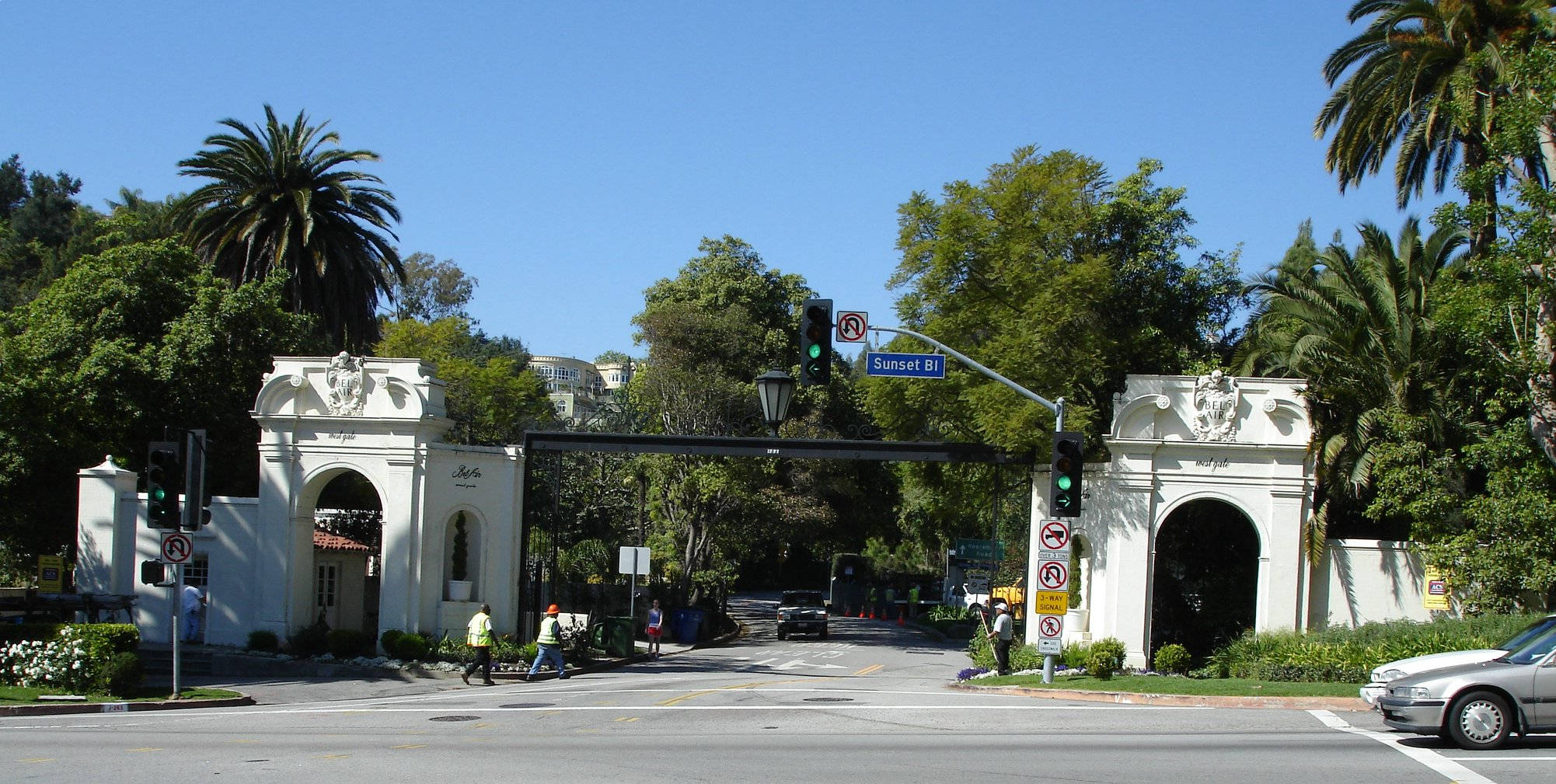 Boulevard 桌布 壁紙 — 一個宏偉的白色拱門，兩旁種有棕櫚樹，框住了晴朗藍天下一個陽光明媚的街道交叉口，有行人和汽車。