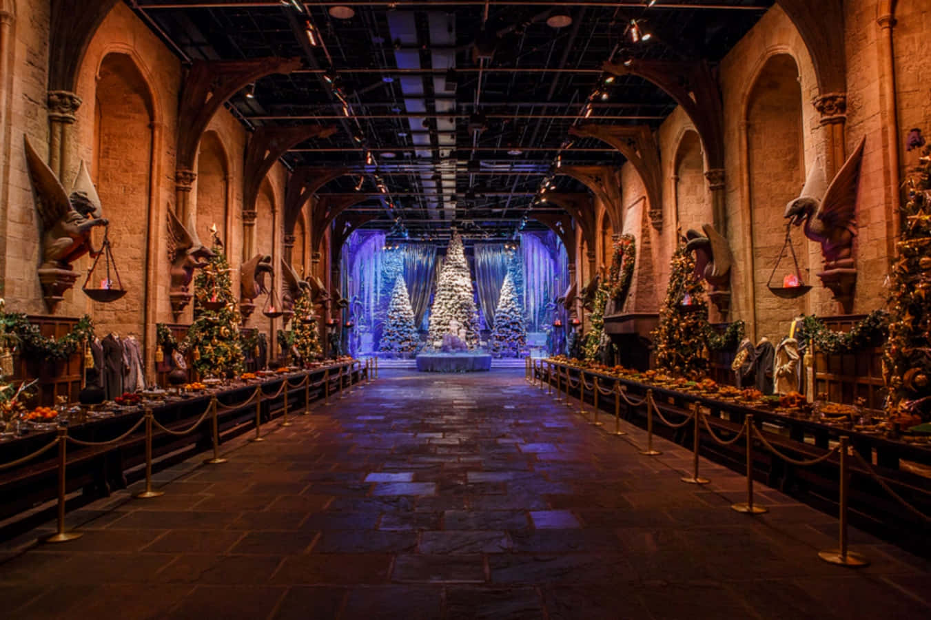 The Hogwarts Great Hall 桌布 壁紙 — 一個宏偉的石廳，在溫暖的環境燈光下裝飾著節日聖誕樹和石像鬼雕像。