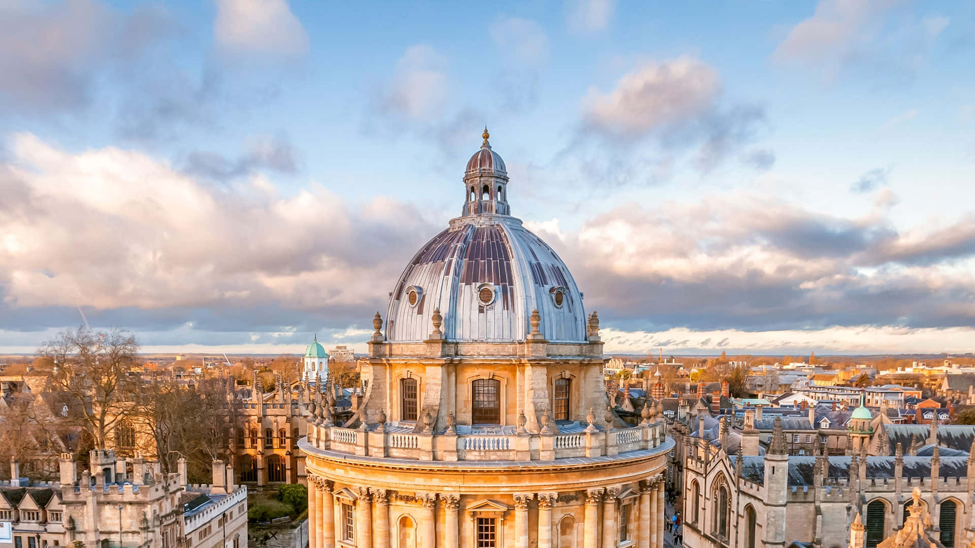 England 桌布 壁紙 — 一個宏偉的石造圓頂配上藍色瓦片屋頂，在柔和的金色日落天空下，矗立於歷史悠久的大學建築之上。