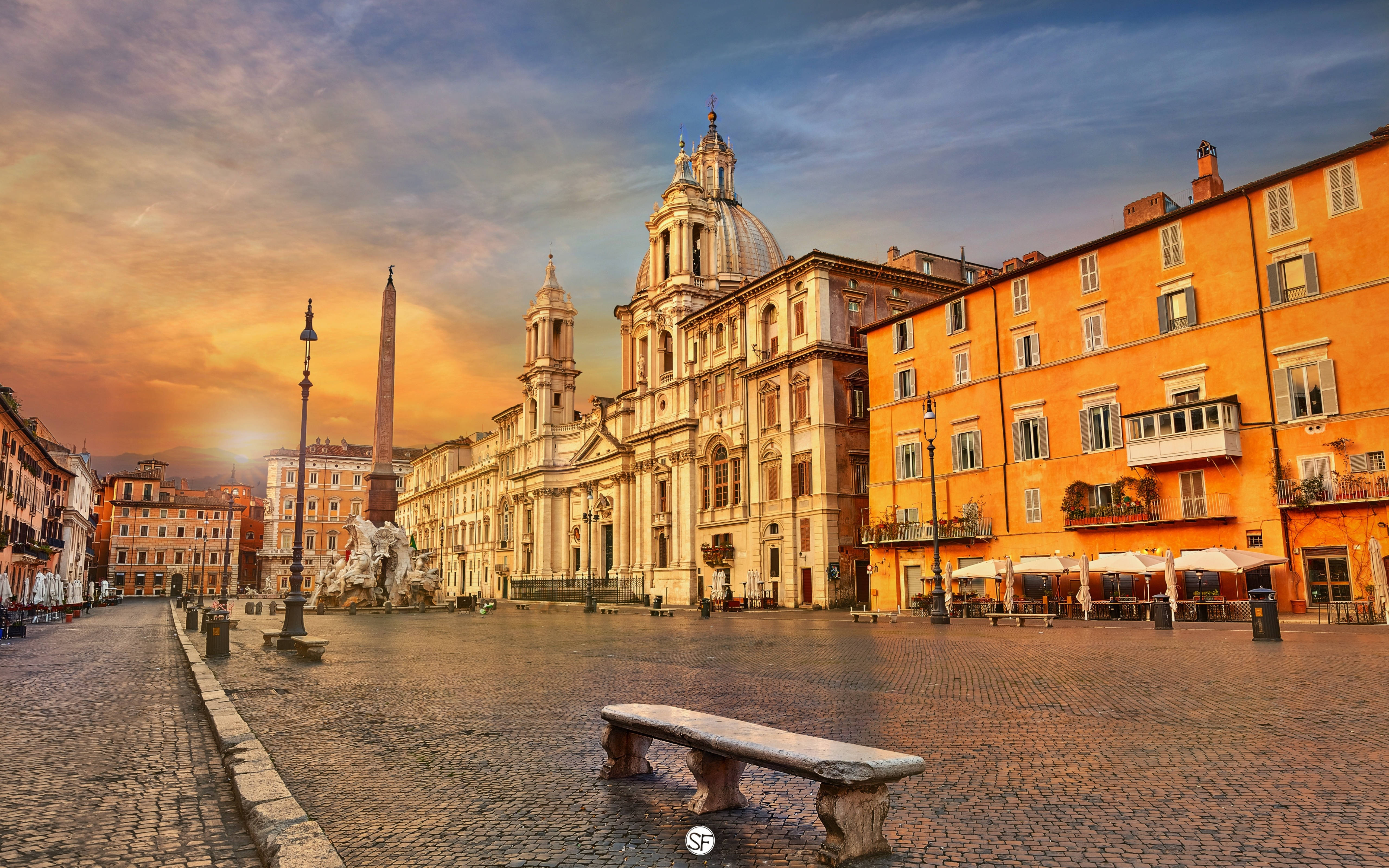 Rome 桌布 壁紙 — 一個宏偉的羅馬廣場，沐浴在金黃色的日落光線中，前景有歷史建築和石凳。