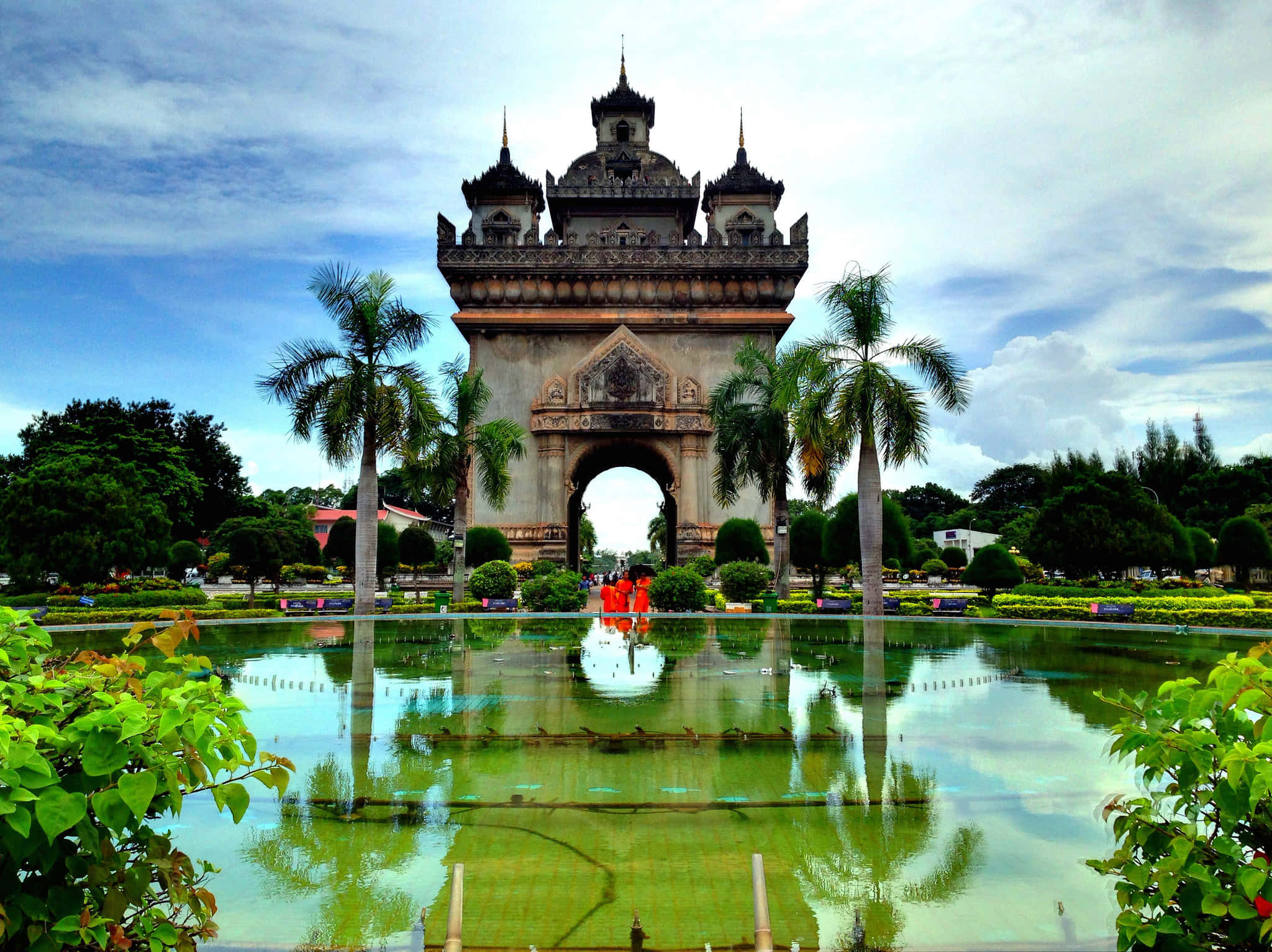 Laos 桌布 壁紙 — 一個宏偉的裝飾拱門帶有尖塔，倒映在一個寧靜的綠色池塘中，周圍環繞著棕櫚樹和茂密的樹葉，天空部分多雲。