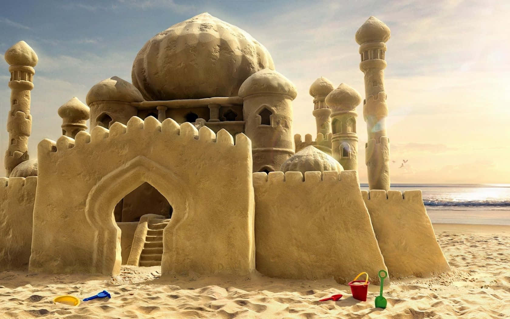 Sandcastles 桌布 壁紙 — 一個宏偉而複雜的沙堡，帶有圓頂和塔樓，矗立在陽光明媚的海灘上，附近有海浪和散落的彩色玩具。