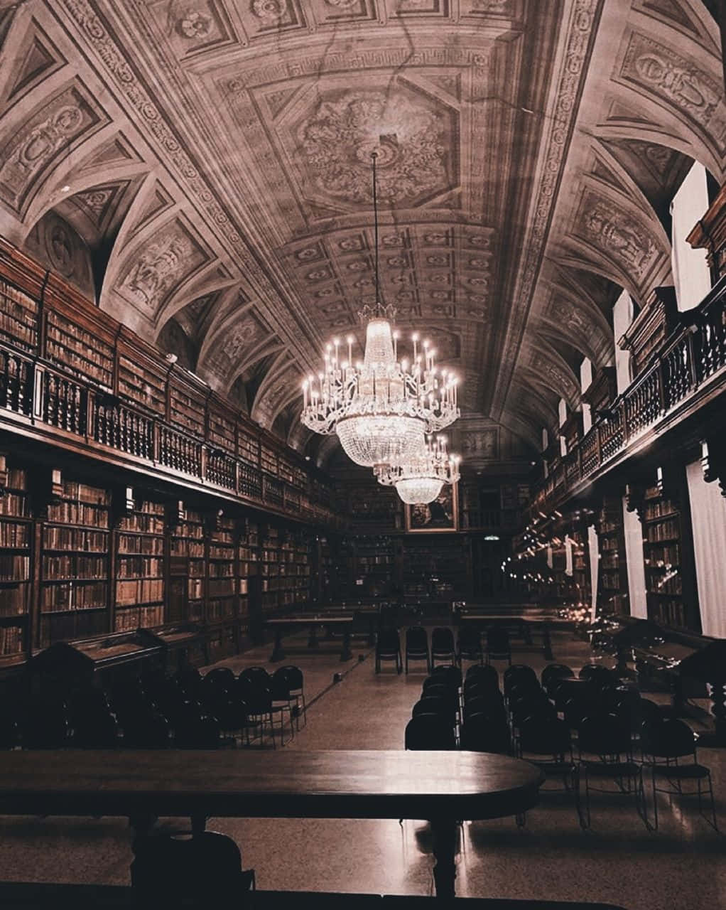 Dark Academia Aesthetic 桌布 壁紙 — 一個宏偉華麗的圖書館，擁有高聳的書架、巨大的吊燈，以及拱形天花板下成排的空書桌。