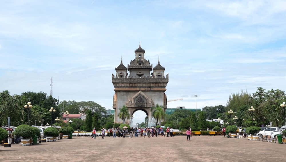 Laos 桌布 壁紙 — 一個宏偉華麗的拱門，帶有複雜的尖塔，在晴朗藍天和零星白雲下，矗立在寬敞的廣場上。