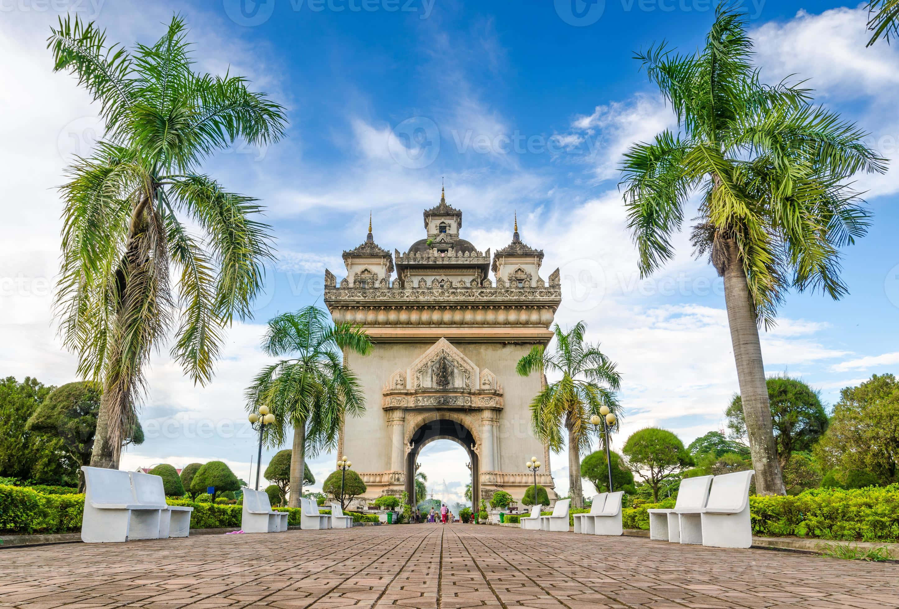 Laos 桌布 壁紙 — 一個宏偉華麗的石拱門矗立在明亮的藍天下，兩旁是高聳的棕櫚樹和白色長椅，位於鋪砌的人行道上。