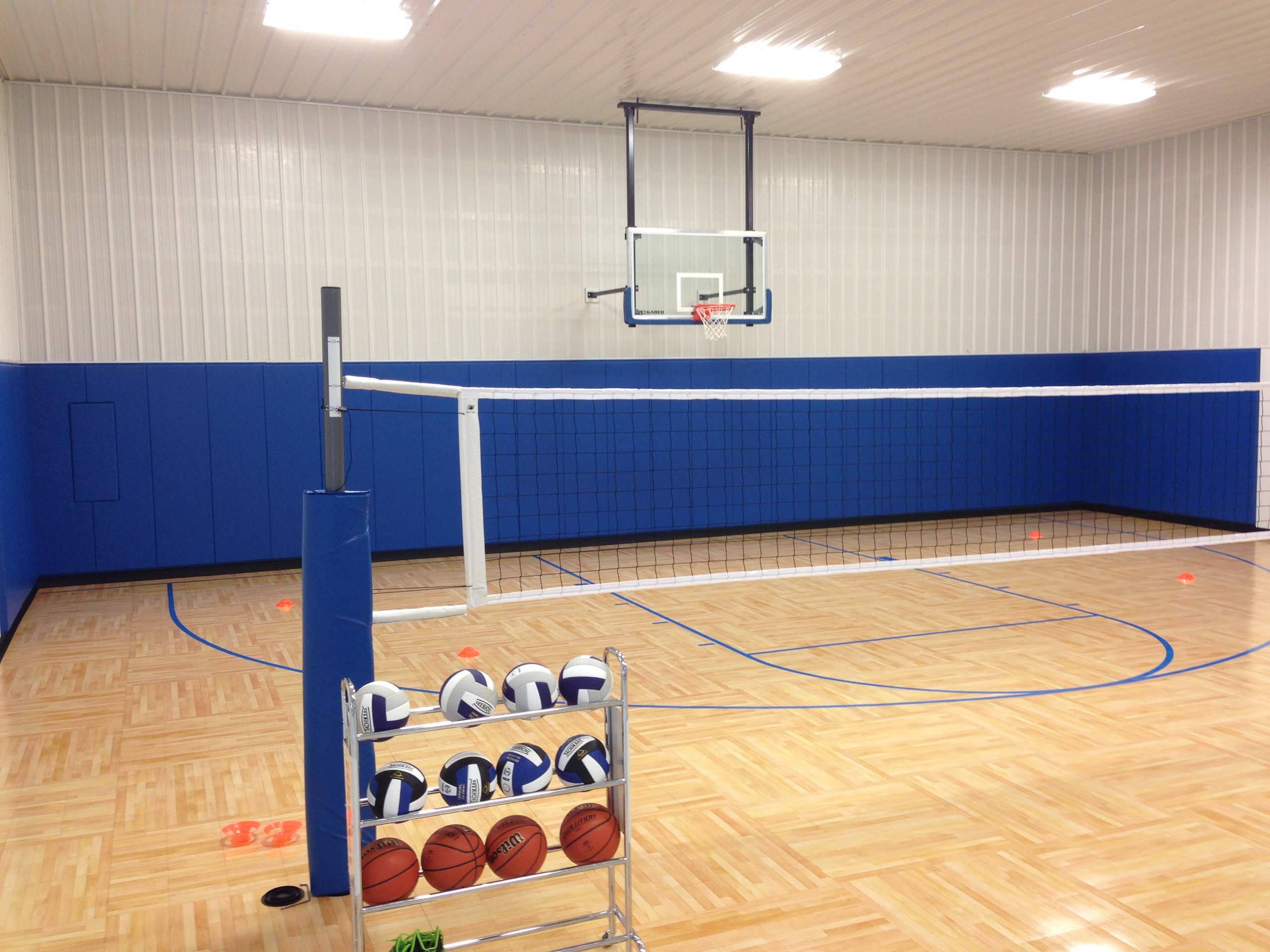 Indoor Volleyball 桌布 壁紙 — 一個室內體育館，設有排球網和籃球框，背景為藍色軟墊牆壁和木地板。
