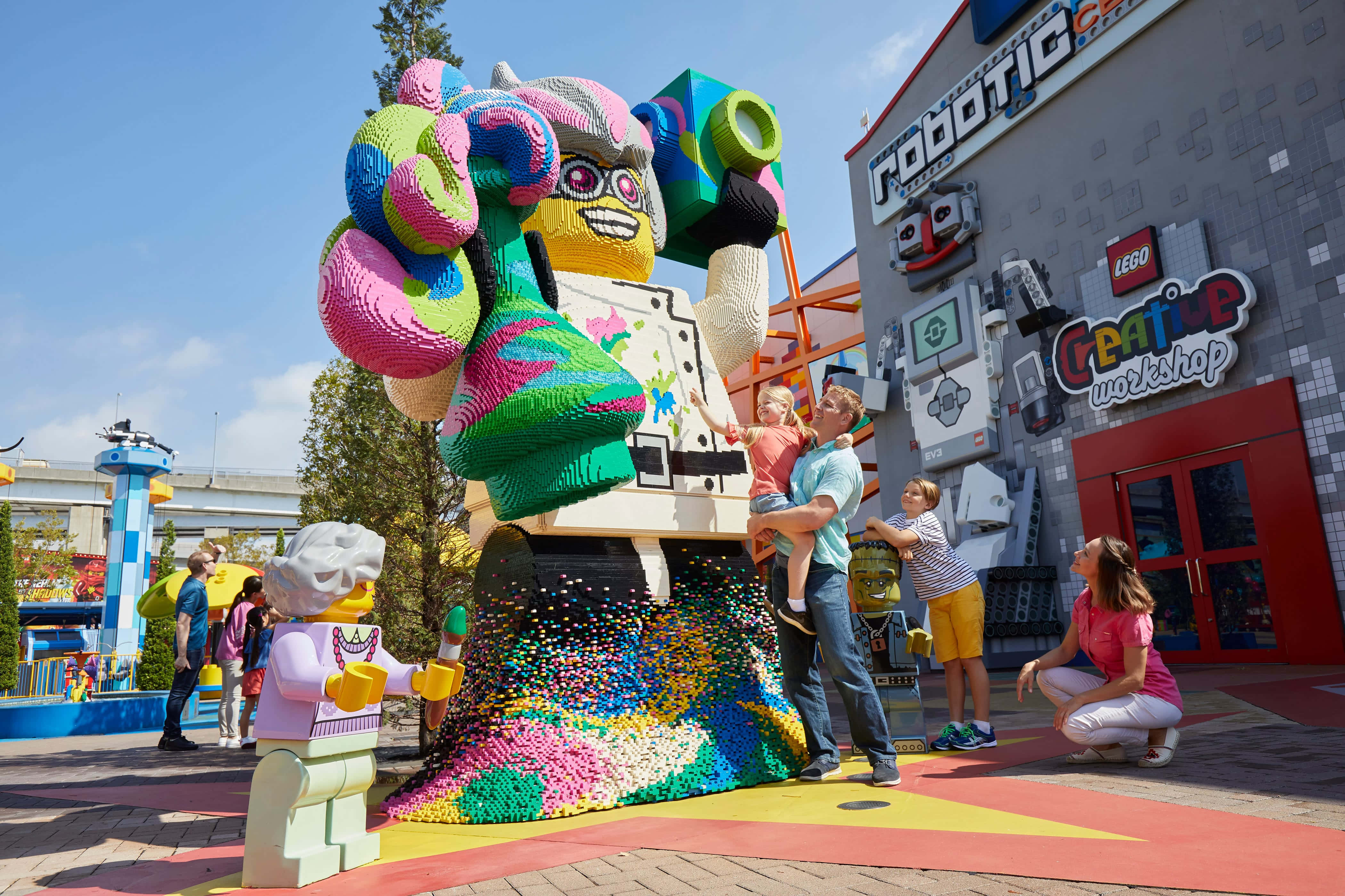 Legoland 桌布 壁紙 — 一個家庭在晴朗藍天下，於充滿活力的主題樂園裡，愉快地在一個巨大的彩色LEGO迷你人偶雕塑旁擺姿勢。