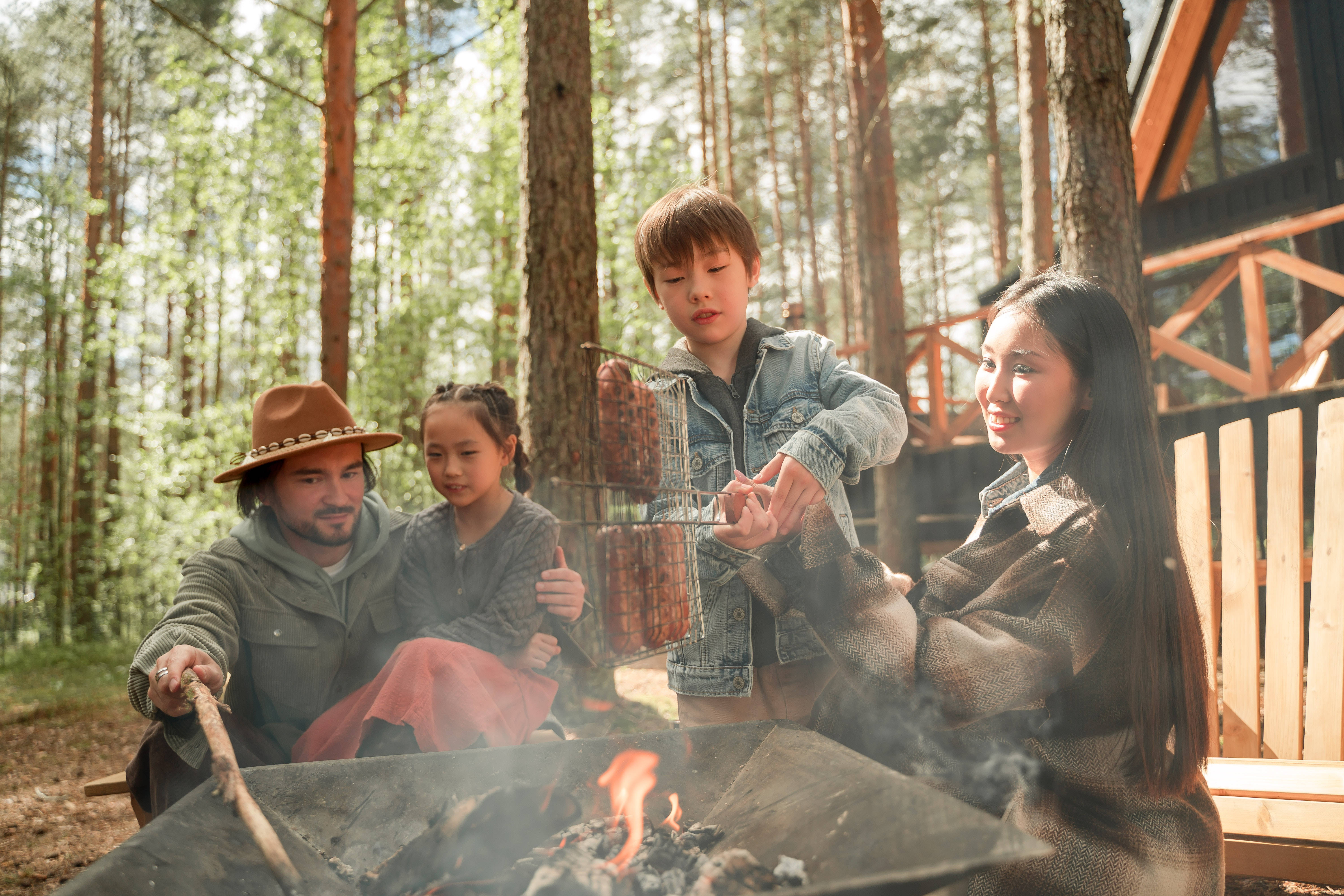 Family 桌布 壁紙 — 一個家庭在陽光普照的松樹林裡，木屋旁邊的營火上烤香腸。