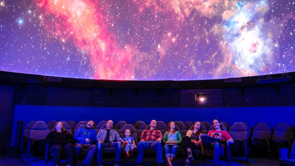 Planetarium 桌布 壁紙 — 一個家庭在黑暗的天文館穹頂下，敬畏地坐在粉紅色、藍色和白色星雲的鮮明宇宙投影之下。