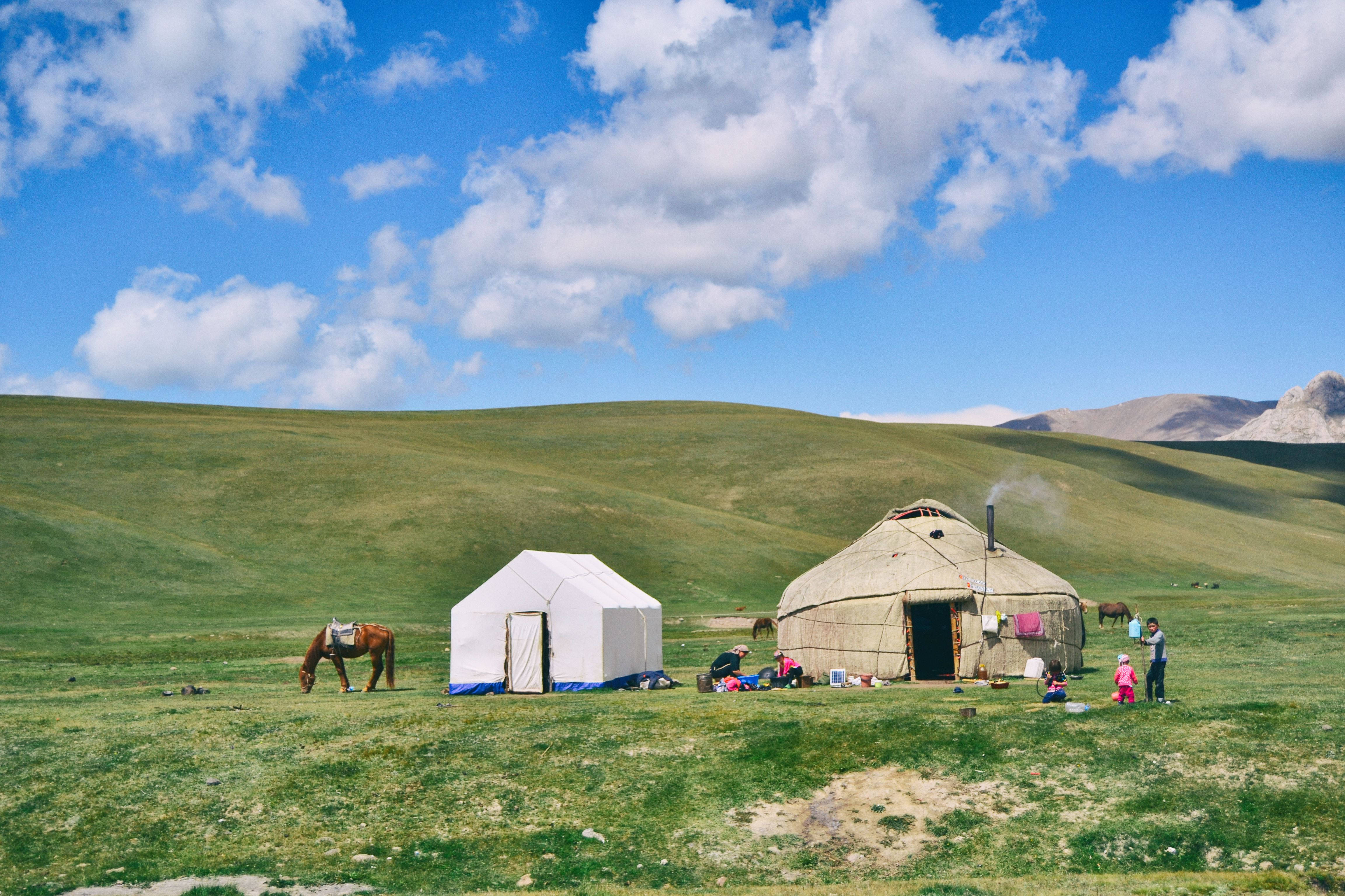 Kyrgyzstan 桌布 壁紙 — 一個家庭聚集在傳統蒙古包和現代帳篷附近，位於廣闊的綠色草原上，天空湛藍，白雲朵朵。