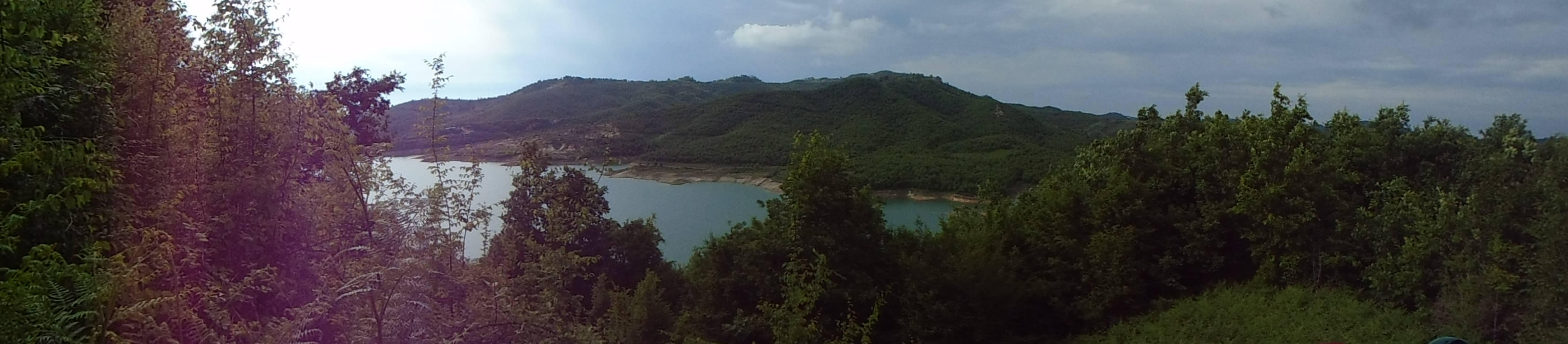Albania 桌布 壁紙 — 一個寧靜湖泊的全景，坐落在鬱鬱蔥蔥的綠色山丘之間，天空多雲，帶有鏡頭光暈。