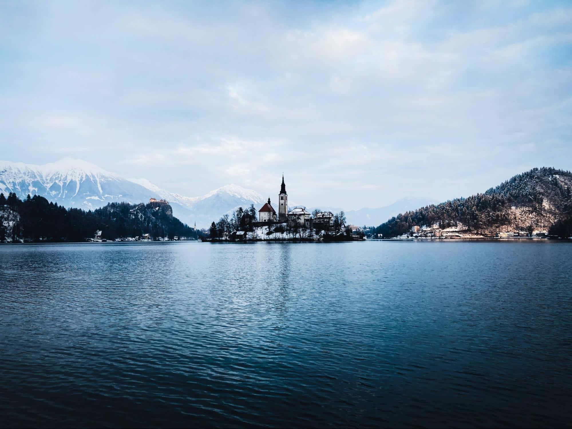 Alps 桌布 壁紙 — 一個寧靜的冬日湖景，有一座被雪覆蓋的島嶼教堂，周圍是森林覆蓋的山丘和遠處白雪覆蓋的山脈，在柔和的藍天下。