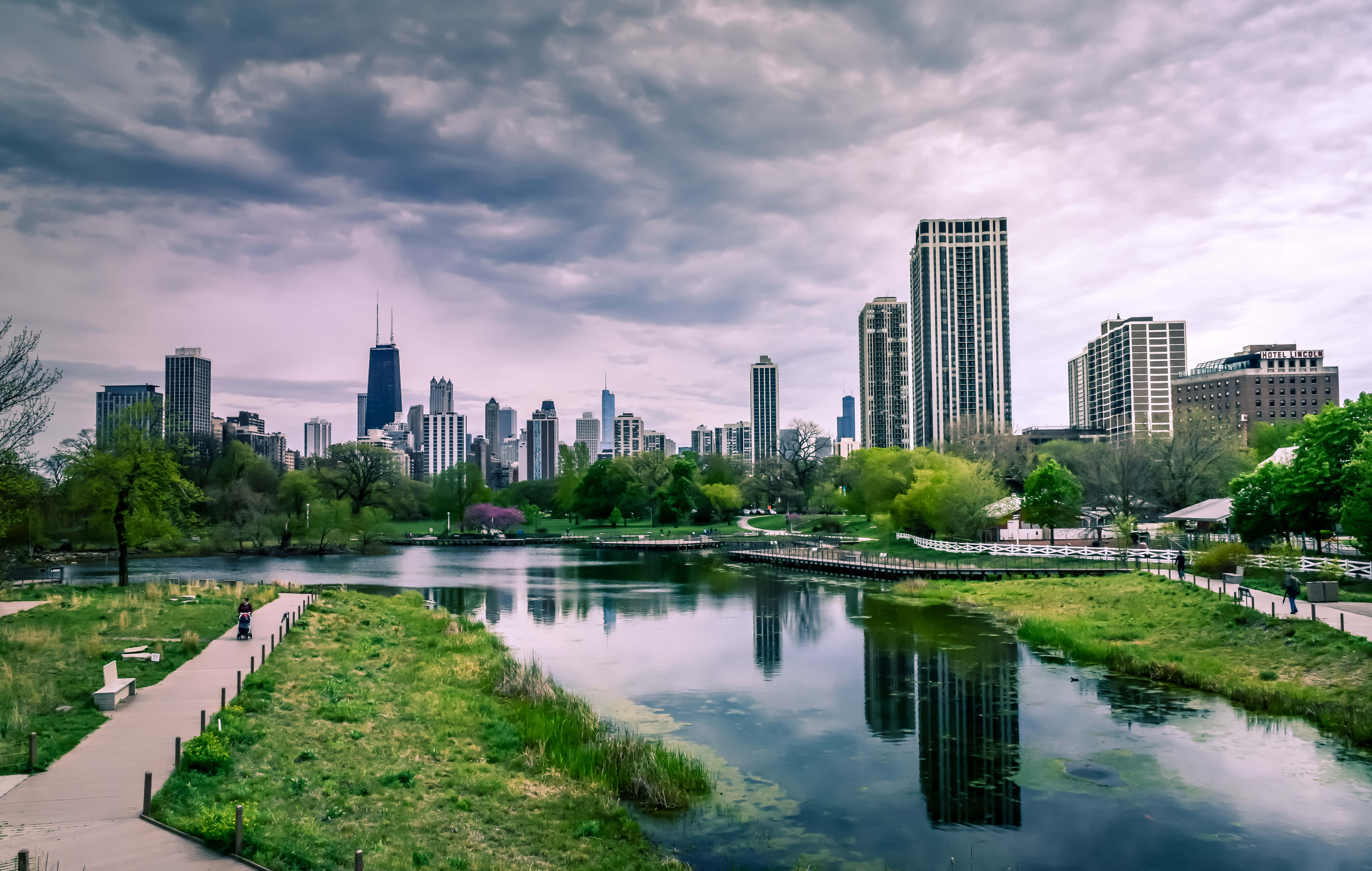 城市風景 桌布 壁紙 — 一個寧靜的城市公園，平靜的湖面倒映著陰沉多雲天空下的高聳摩天大樓。