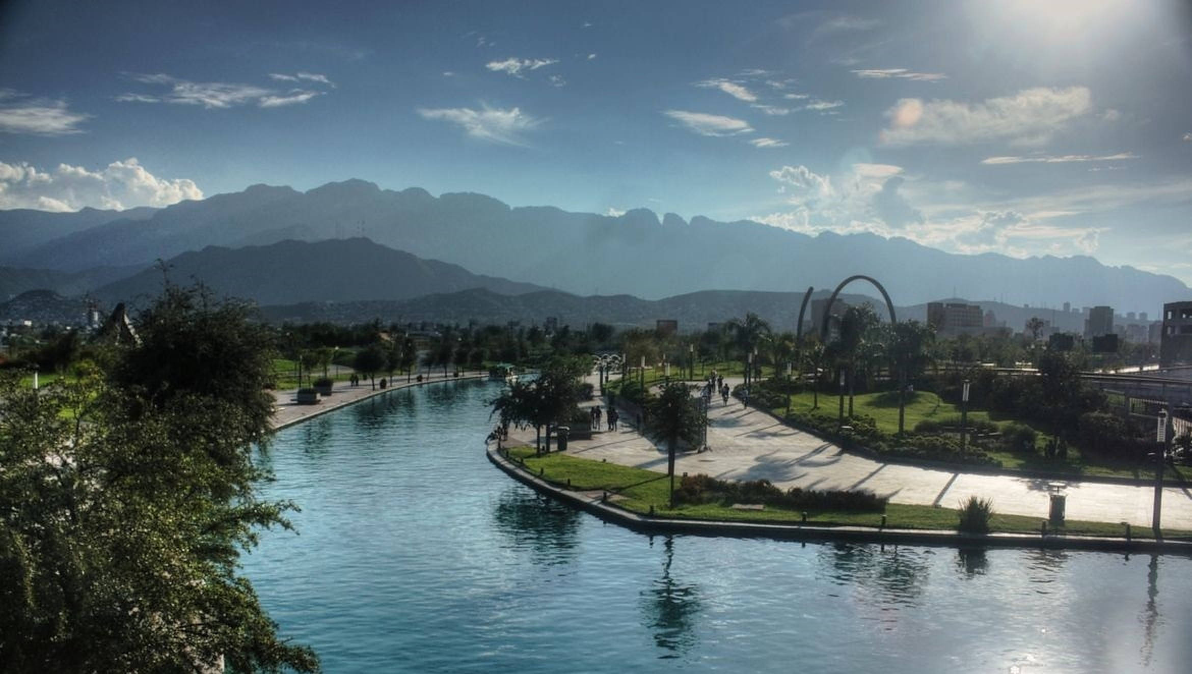 Monterrey 桌布 壁紙 — 一個寧靜的城市公園，蜿蜒的運河兩旁是步道和棕櫚樹，頭頂是明亮的藍天，遠處有山巒。