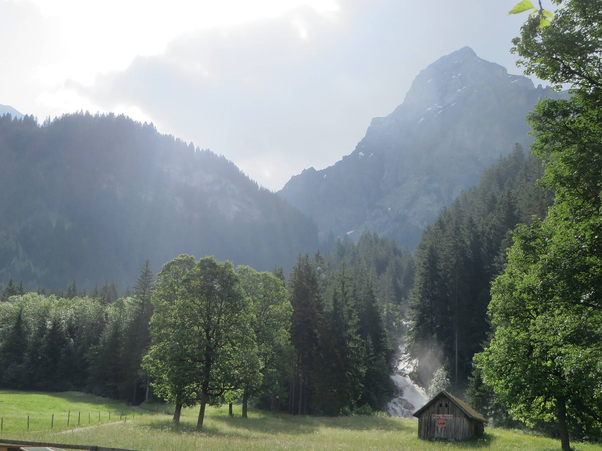 Alps 桌布 壁紙 — 一個寧靜的山谷，木屋坐落在茂密的綠樹叢中，霧氣繚繞的瀑布沿著森林覆蓋的山坡傾瀉而下。