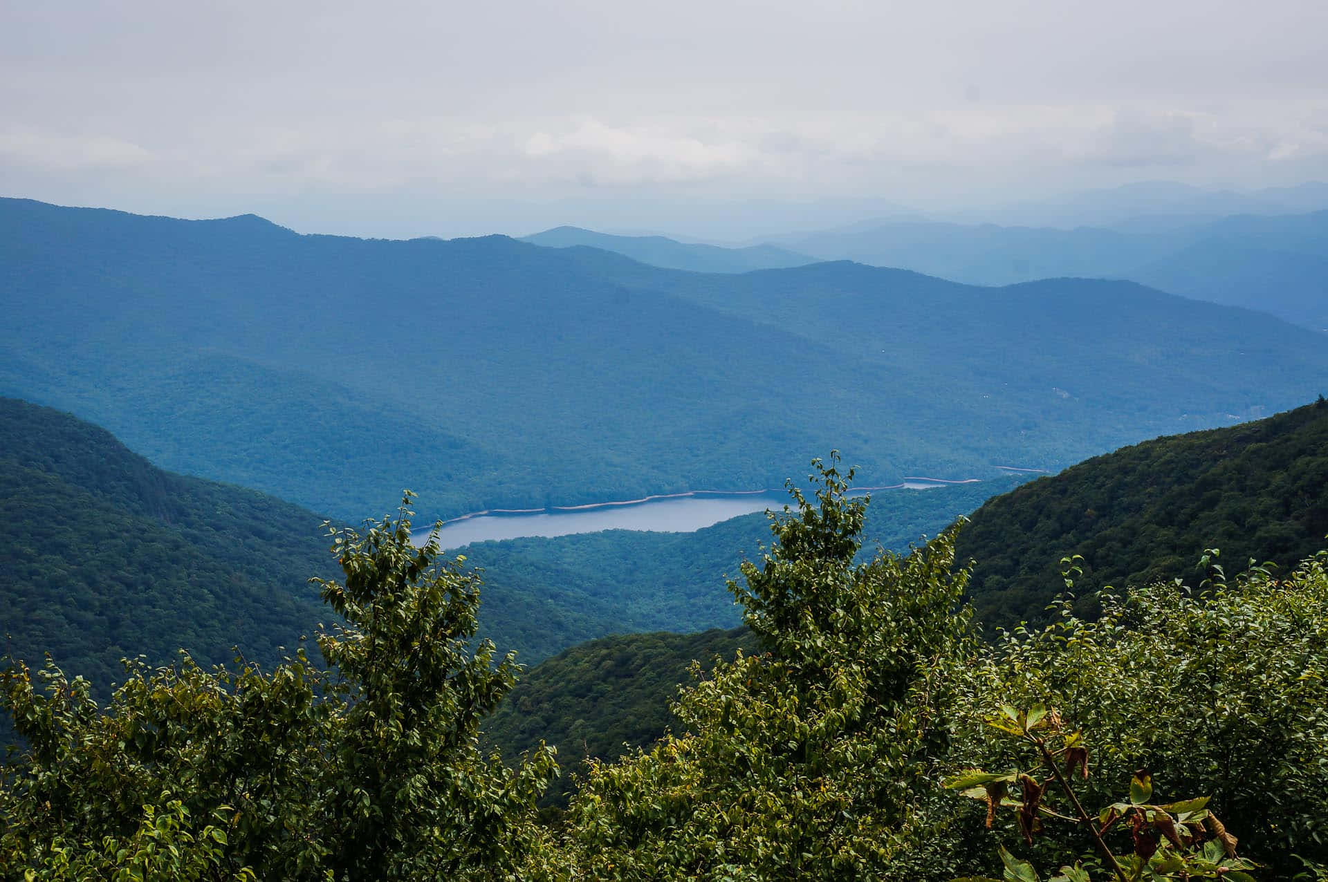 Blue Ridge Parkway 桌布 壁紙 — 一個寧靜的山谷，蜿蜒的湖泊坐落在層層疊疊的森林山丘之間，天空多雲。
