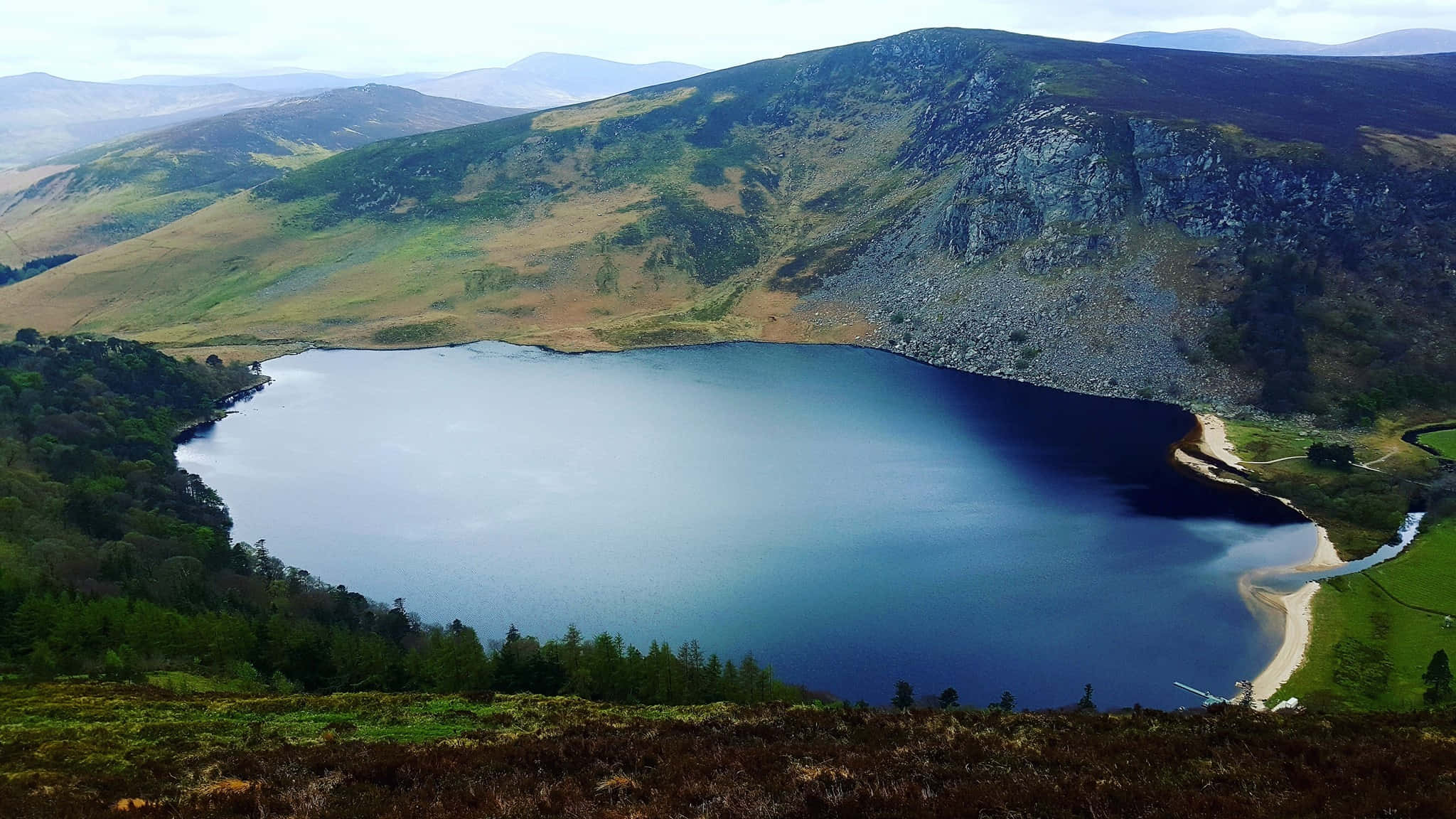 Lake 桌布 壁紙 — 一個寧靜的山間湖泊，坐落在崎嶇的綠色山丘中，天空柔和多雲。